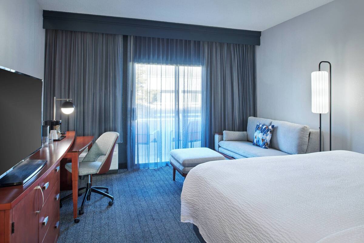 A hotel room with a bed, a desk, a chair, a sofa, and a large window with curtains.