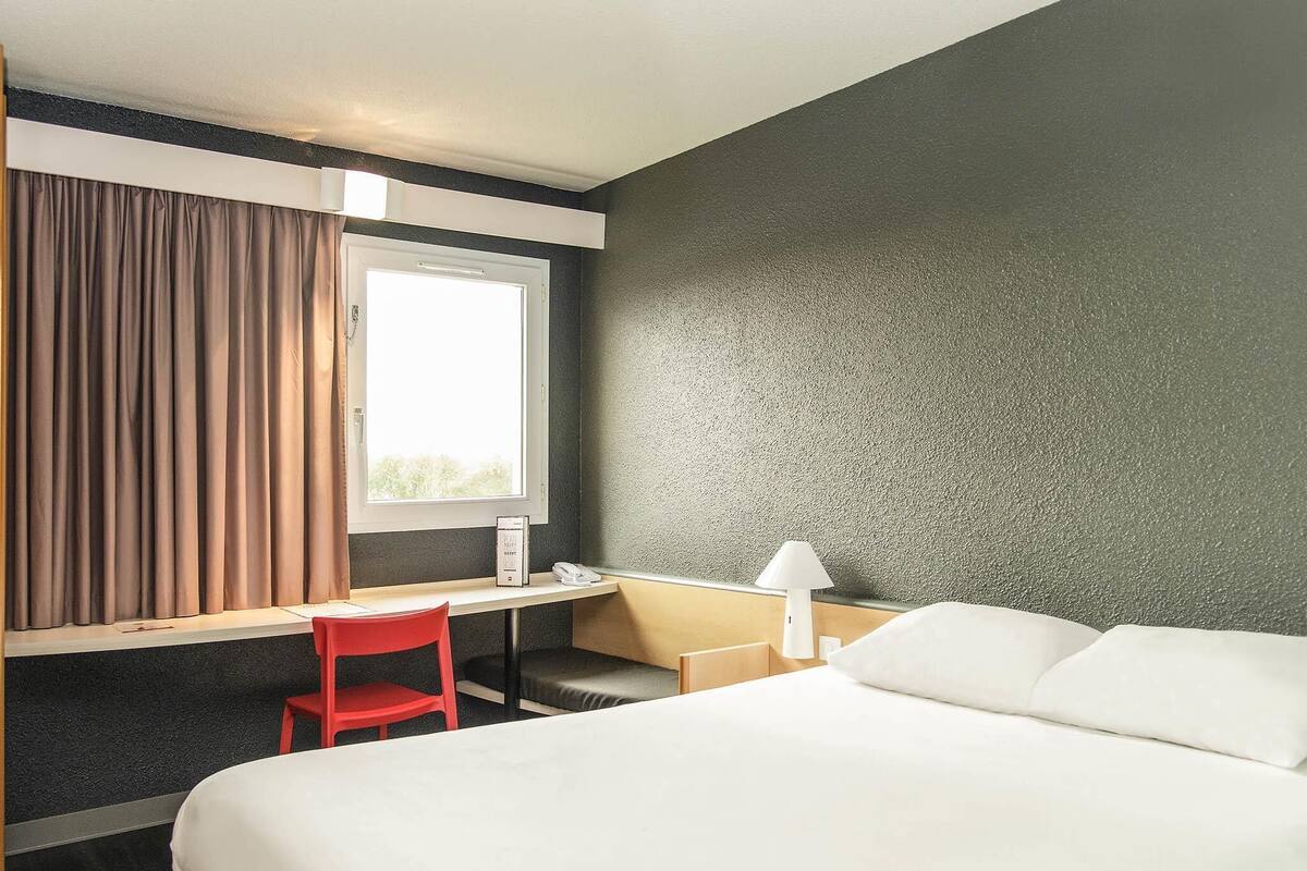 A hotel room with a bed, a desk, a chair, a lamp, and a window with curtains.