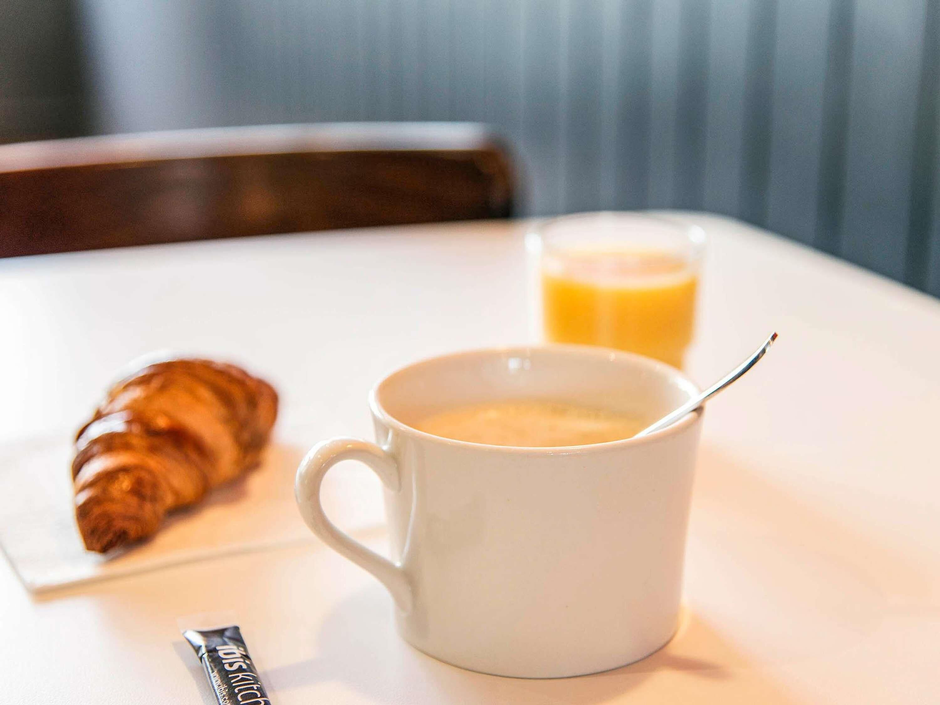 Une tasse blanche avec une cuillère, un verre de jus d’orange et un croissant sur une table blanche.