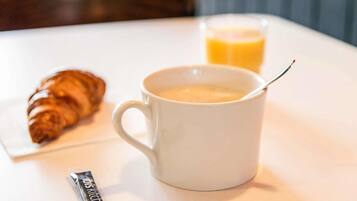Une tasse blanche avec une cuillère, un verre de jus d’orange et un croissant sur une table blanche.