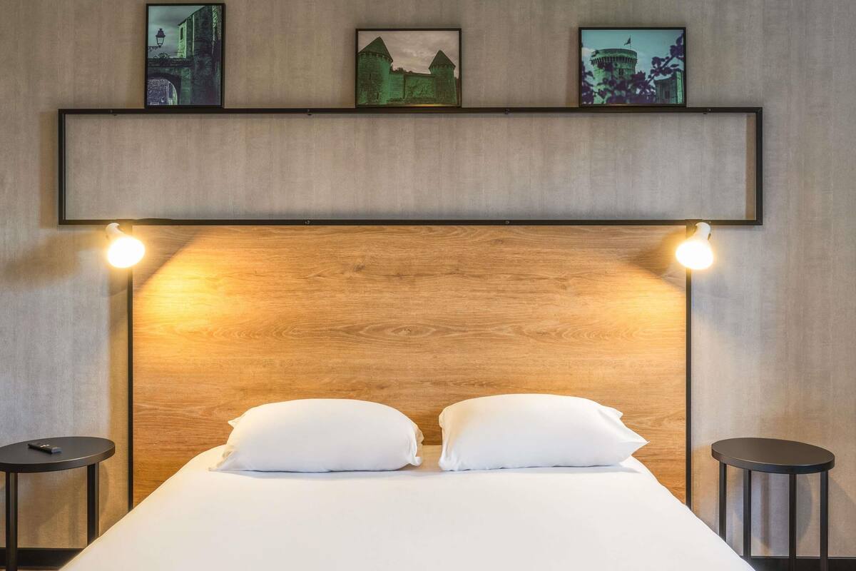 Une chambre d’hôtel avec une tête de lit en bois, deux tables de chevet, un lit avec une literie blanche et trois tableaux encadrés accrochés au mur.