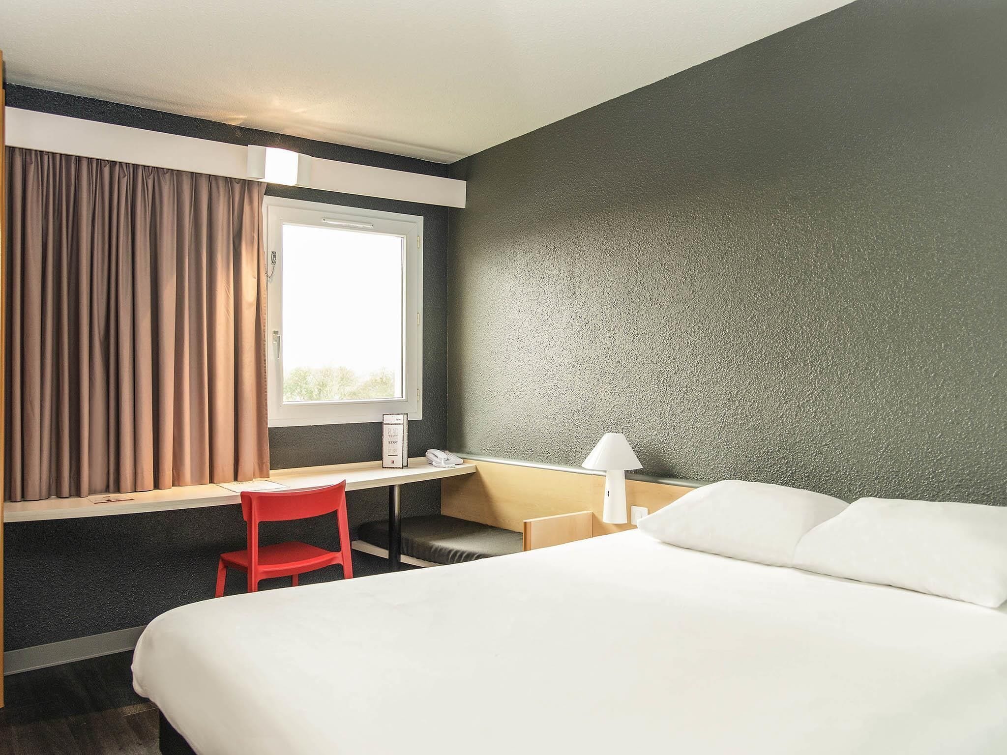 A hotel room with a bed, a desk, a chair, a lamp, and a window with curtains.