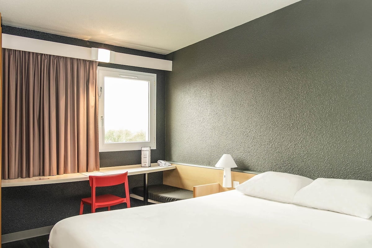 A hotel room with a bed, a desk, a chair, a lamp, and a window with curtains.