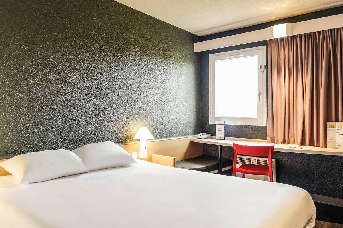 Ein Hotelzimmer mit Bett, Schreibtisch, Stuhl, Lampe und einem fenster mit Vorhängen.
