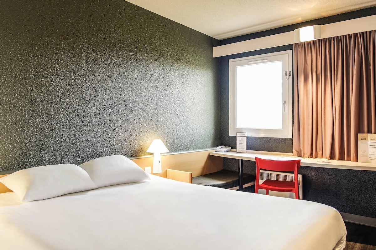 A hotel room with a bed, desk, chair, lamp, and window with curtains.