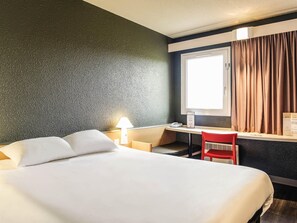A hotel room with a bed, desk, chair, lamp, and window with curtains.