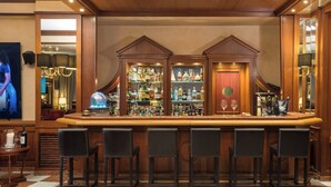 Una zona de bar con una barra de madera, estantes con botellas de licores y varios taburetes negros.