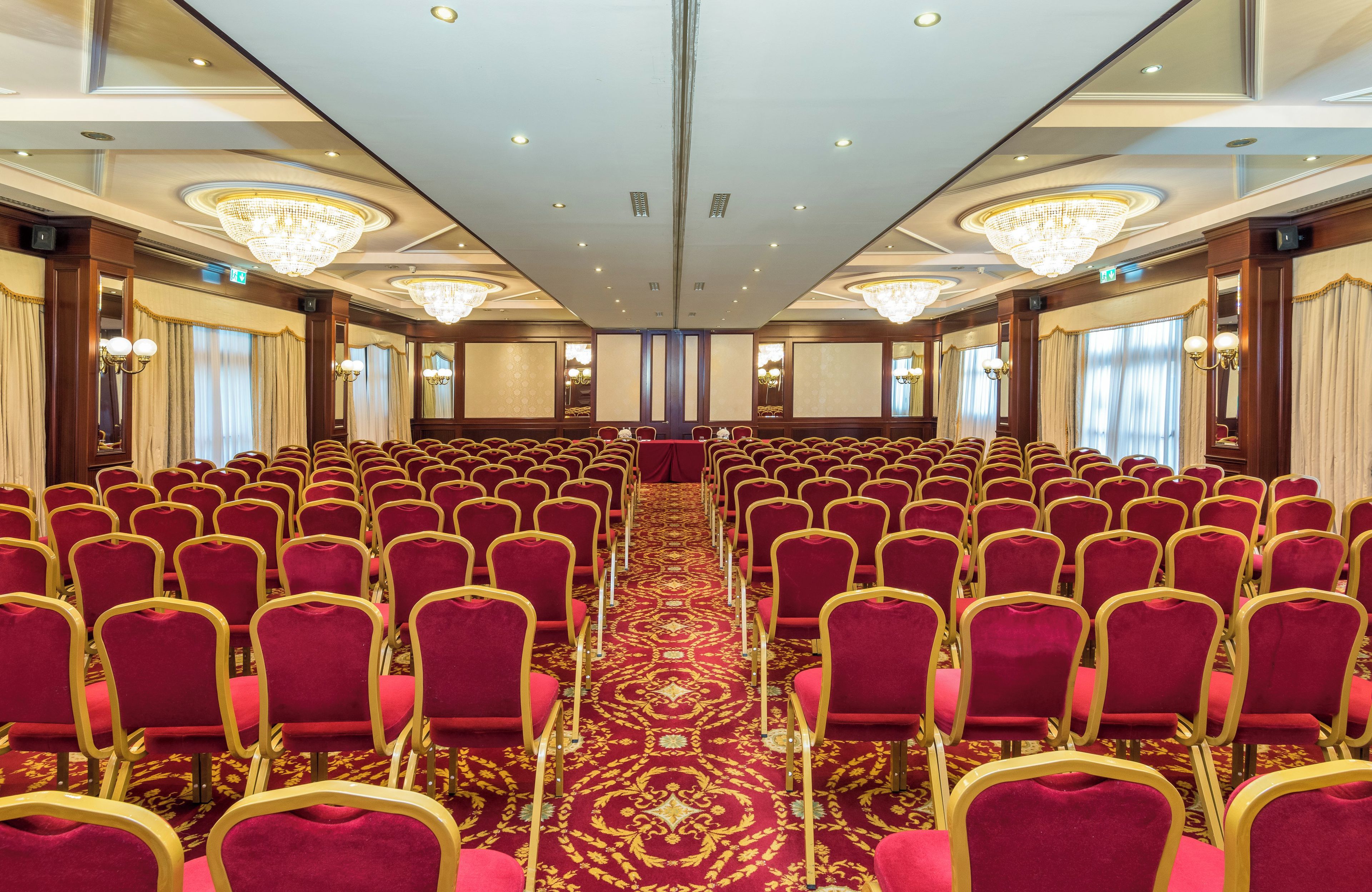 A conference room with rows of red chairs, a patterned carpet, and chandeliers.