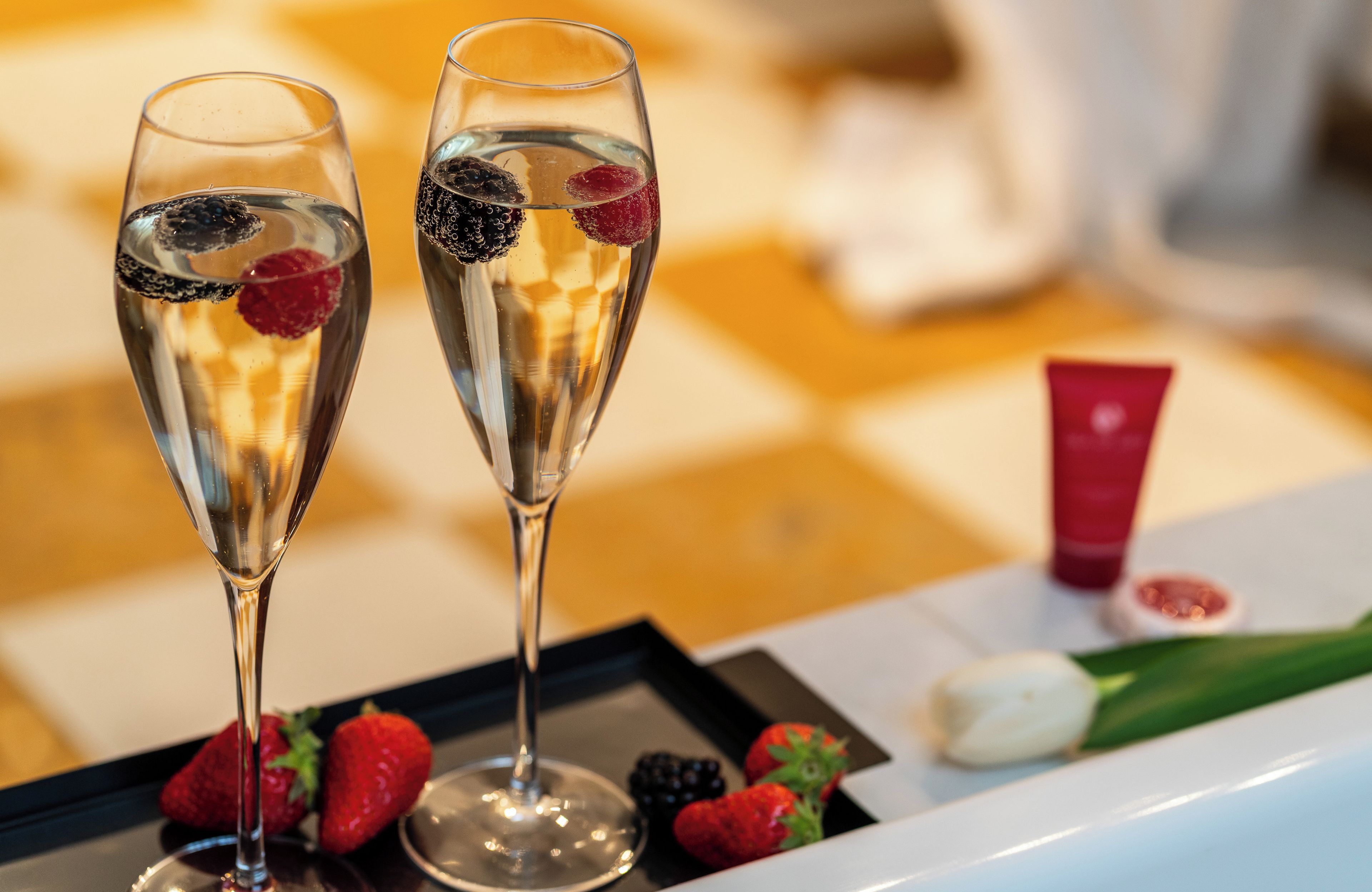 Two champagne flutes with fruit garnishes on a tray, accompanied by strawberries and a red tube, likely a cosmetic product, on a checkered tablecloth.