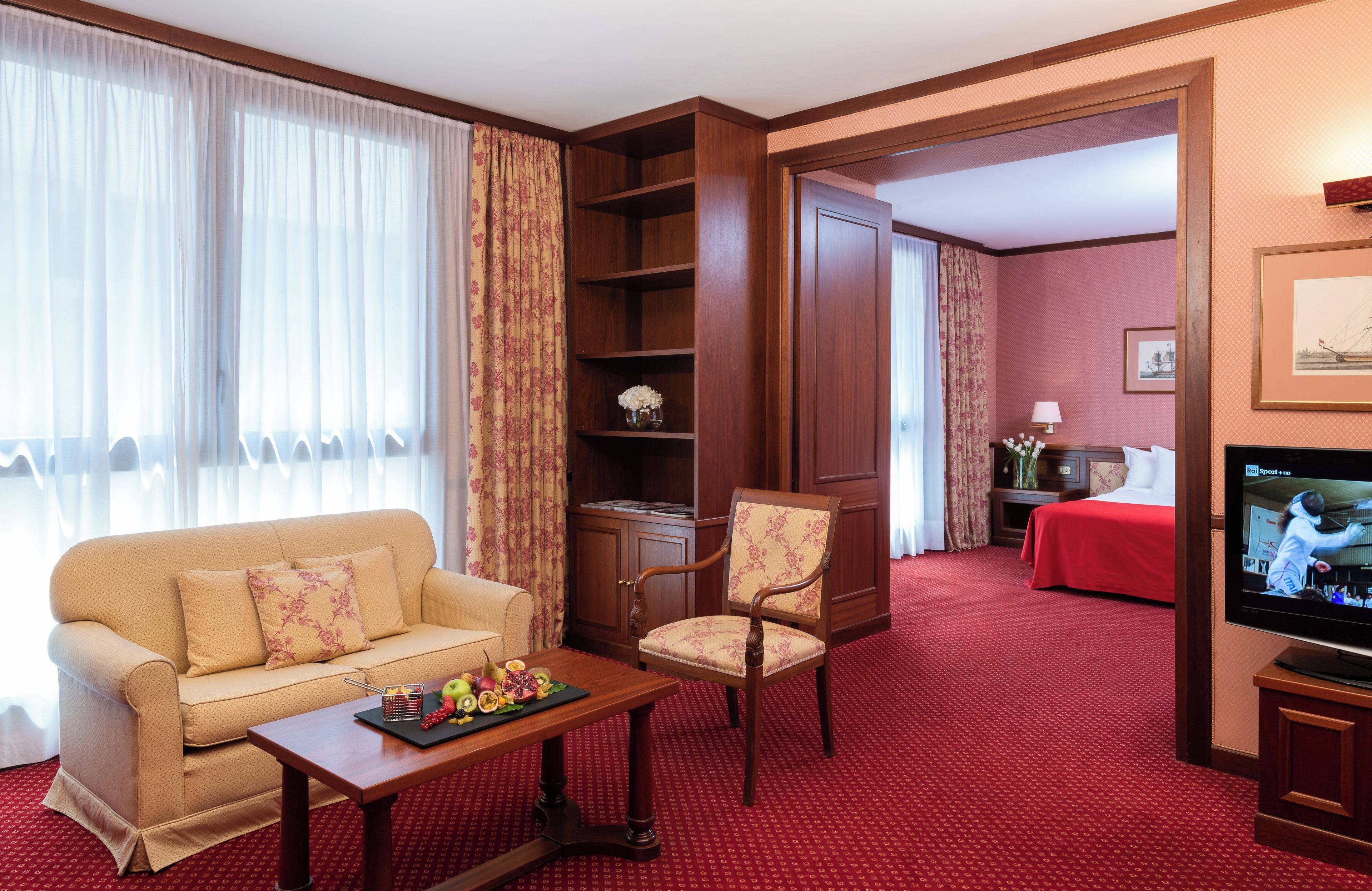 A hotel room with a sofa, armchair, TV, and a red carpet.