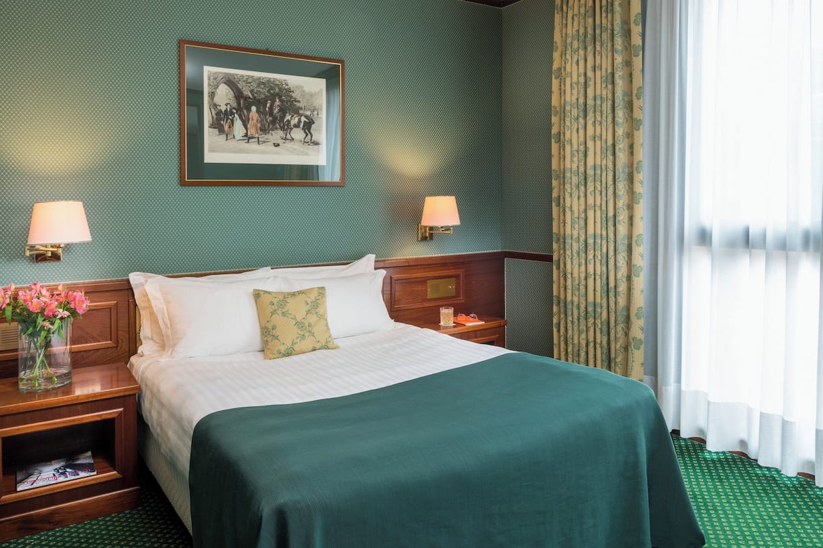 Un lit bien fait, recouvert d’une couverture verte et agrémenté d’un seul coussin décoratif. Deux tables de chevet avec des lampes se trouvent de part et d’autre du lit, et un tableau encadré est accroché au-dessus.