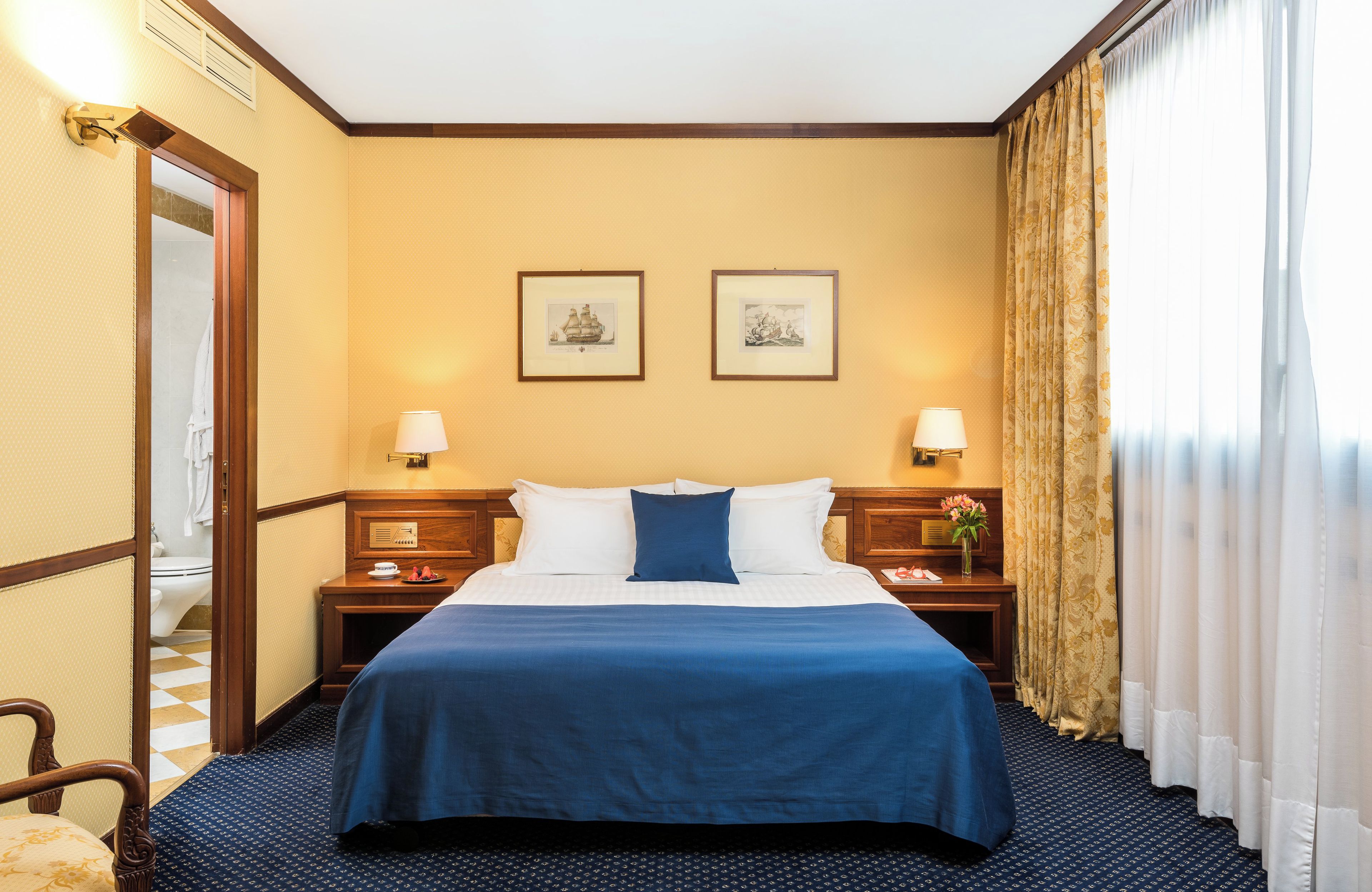 Quarto de hotel com uma cama grande, duas mesas de cabeceira com abajures, uma cadeira de madeira e vista para o exterior através de uma janela com cortinas.