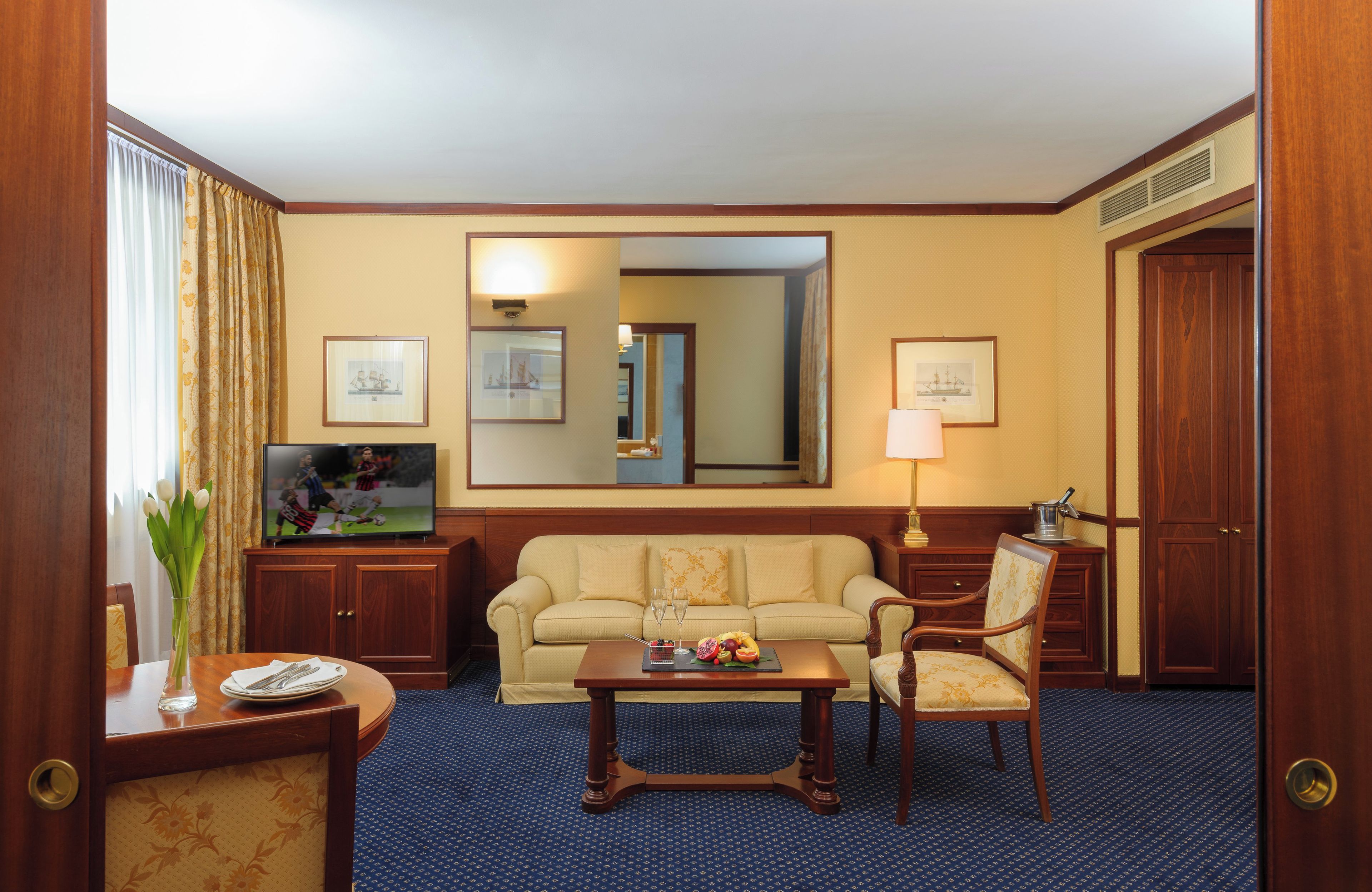 Une chambre d’hôtel équipée d’un canapé, de fauteuils, d’une table basse, d’une télévision, d’un miroir et de tableaux encadrés accrochés au mur.