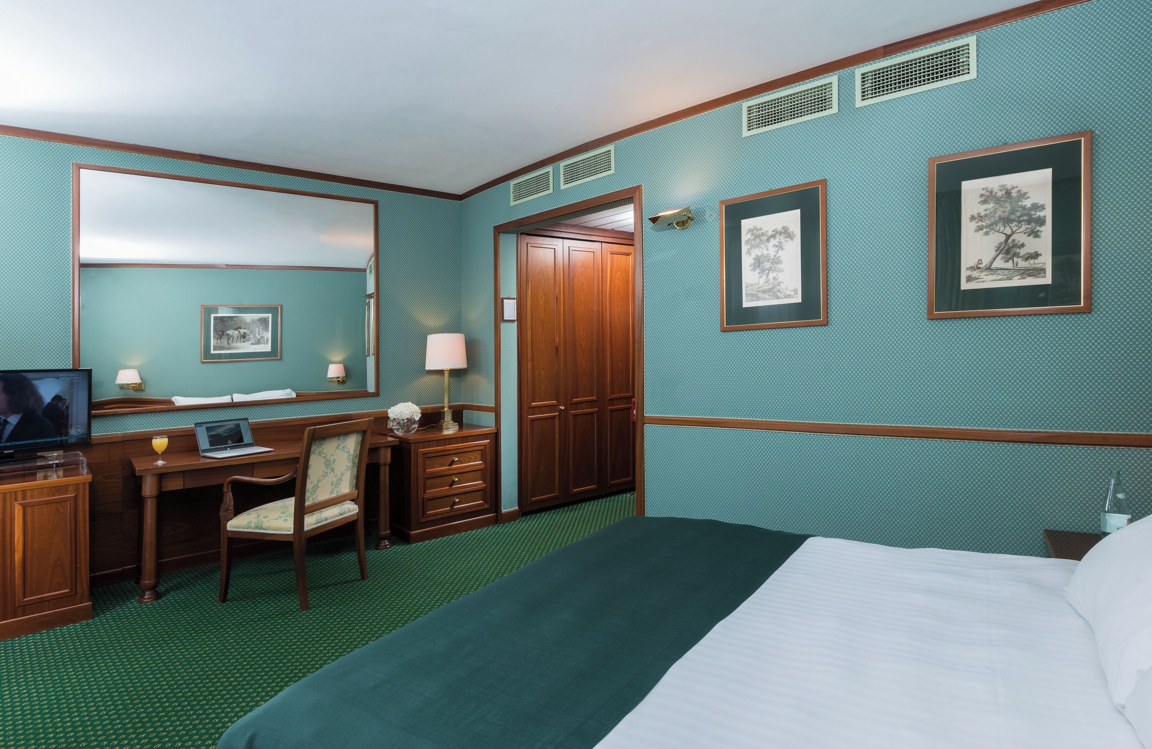 A hotel room with a bed, a desk with a chair, a television, a dresser, and framed pictures on the wall.