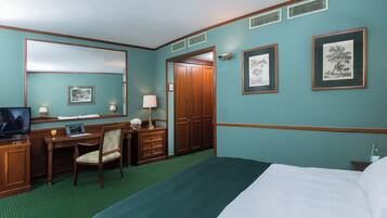A hotel room with a bed, a desk with a chair, a television, a dresser, and framed pictures on the wall.