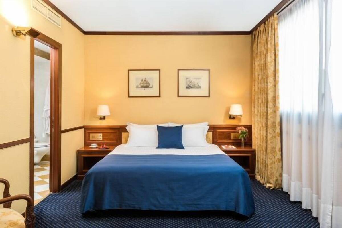 Habitación de hotel con cama, dos mesitas de noche, lámparas, una ventana con cortinas y dos cuadros en la pared.