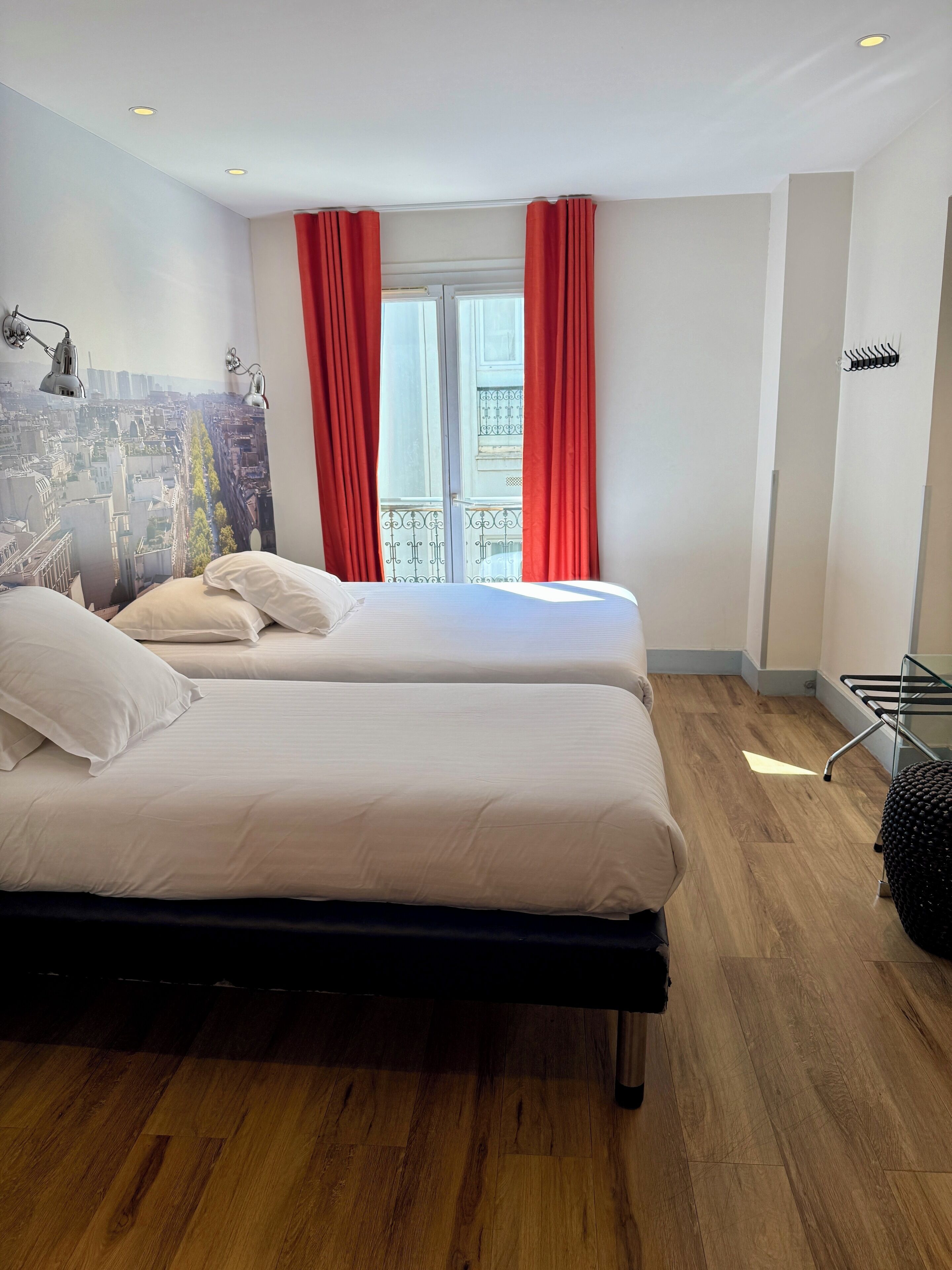 Quarto de hotel com duas camas, um mural com vista urbana na parede, varanda com cortinas vermelhas e piso de madeira.