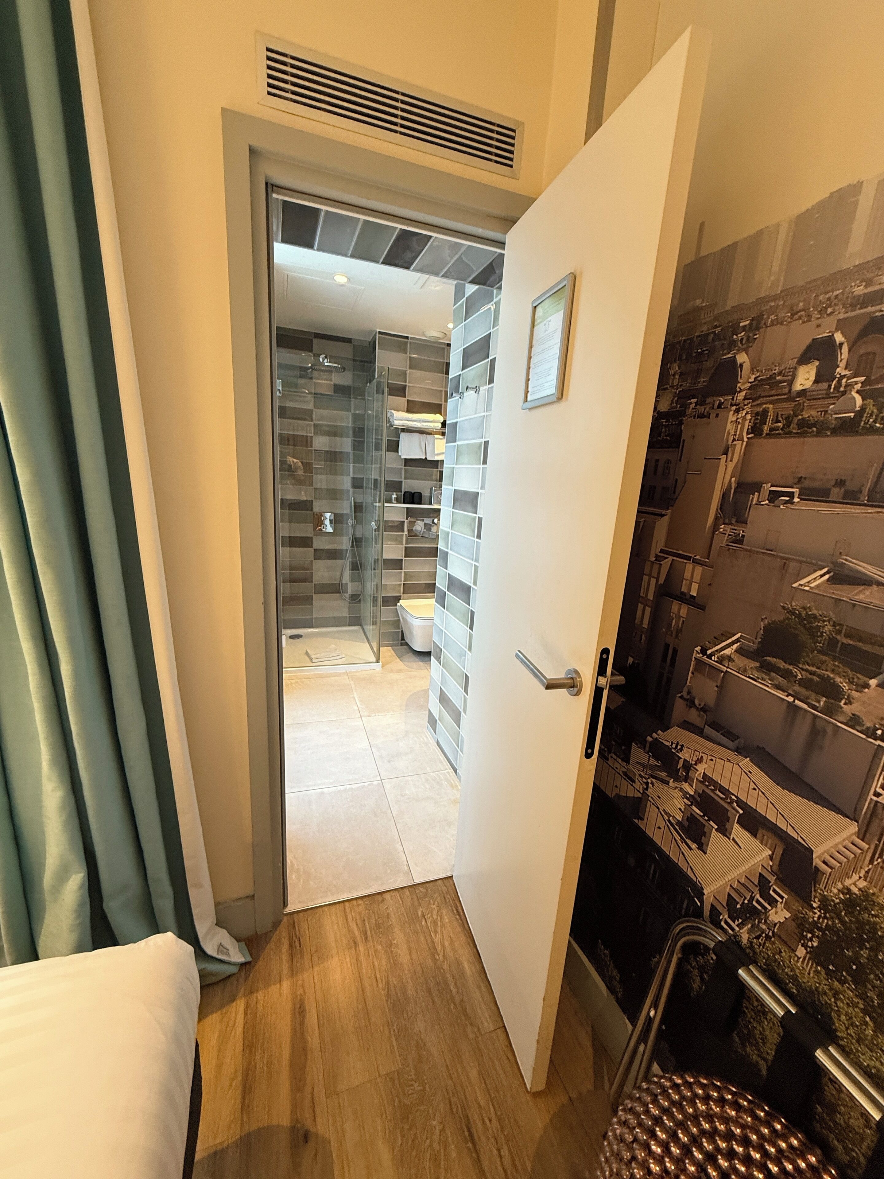A hotel room with a wooden floor, a bed, a bathroom with a glass shower, and a view of buildings through the window.