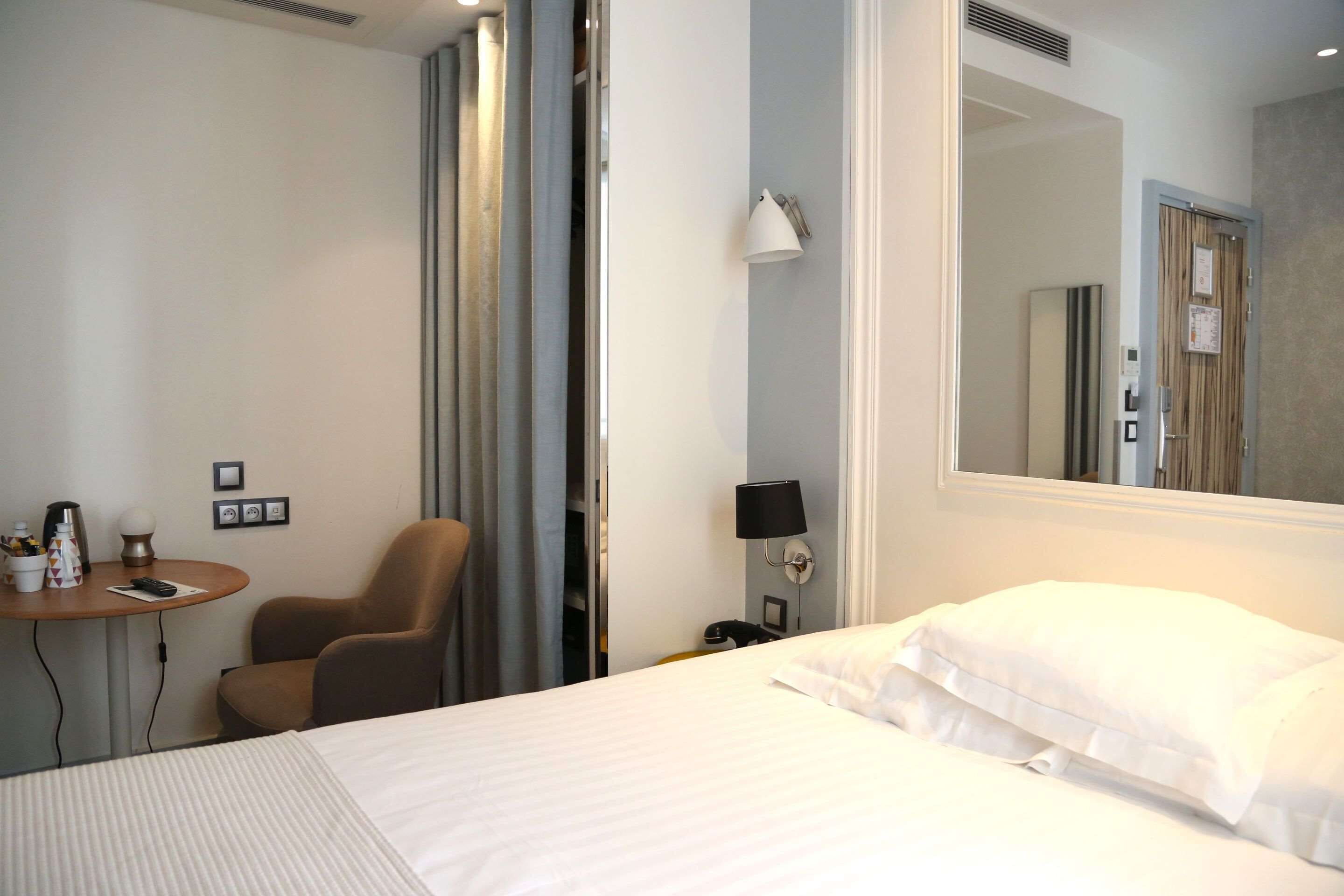 A hotel room with a bed, a chair, a table with a lamp, and a mirror on the wall.