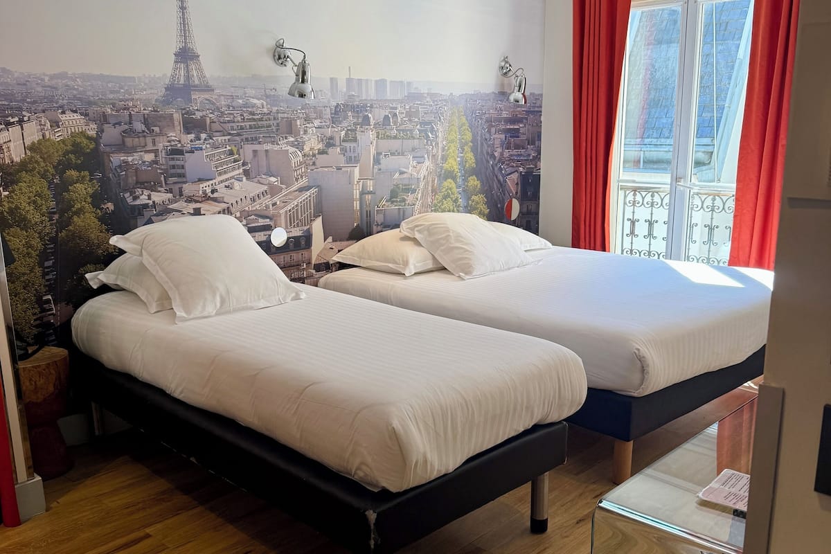 A hotel room with two beds, a cityscape mural, wooden flooring, and a window with red curtains.
