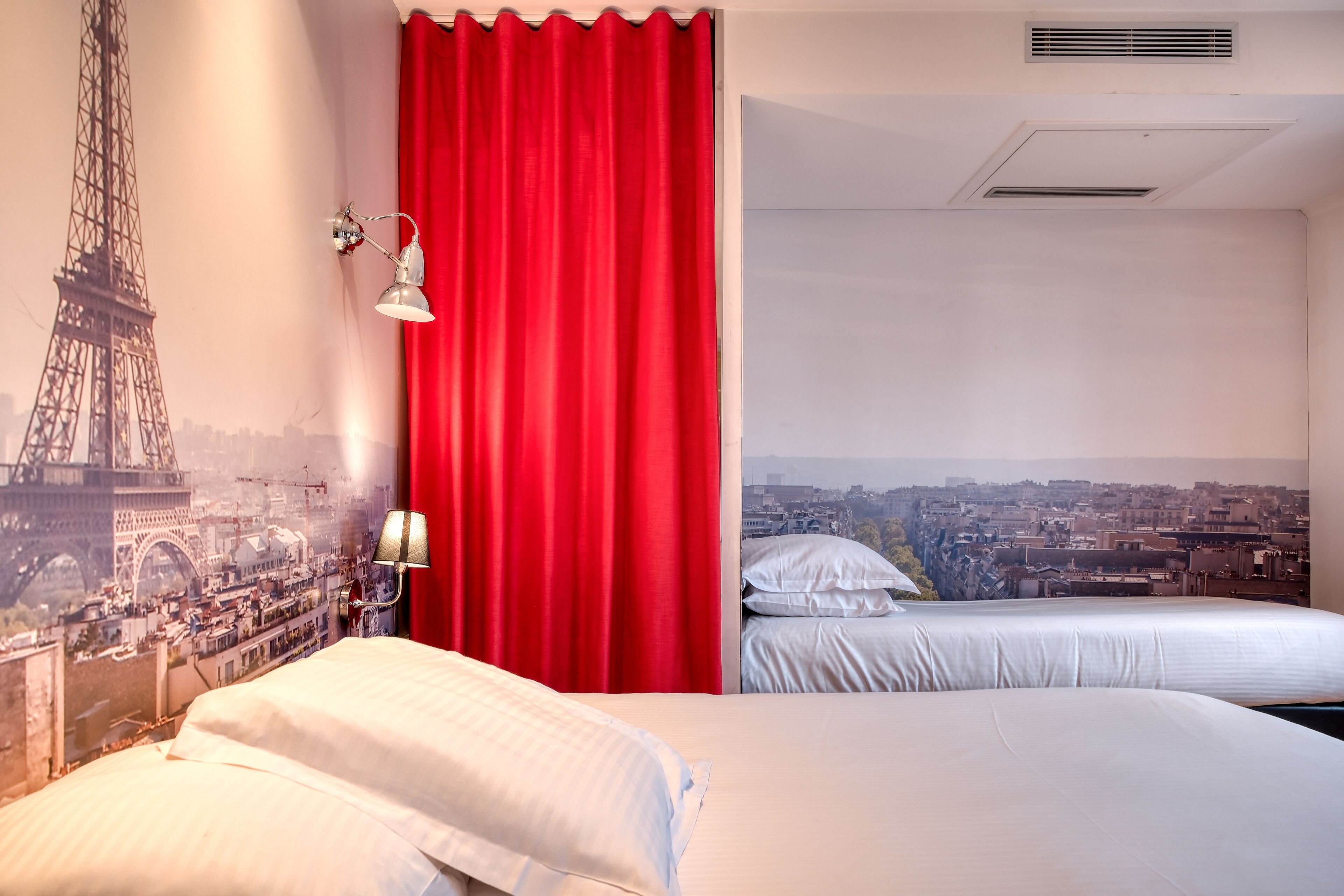 A hotel room with a bed, a red curtain, a wall-mounted lamp, and a view of a cityscape through a window.