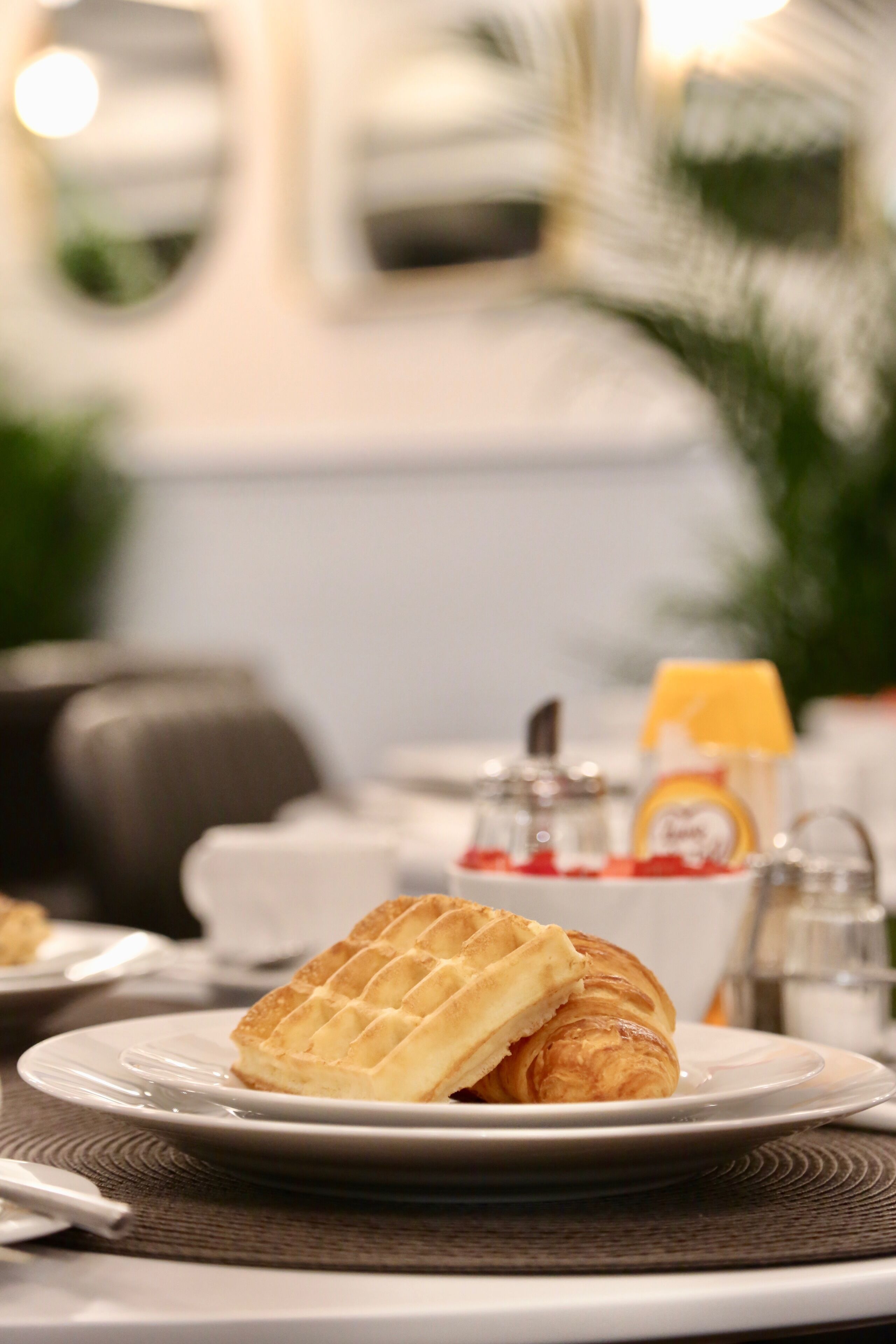 A breakfast setting with a waffle and croissant on a plate, a coffee pot, and a bottle of orange juice.