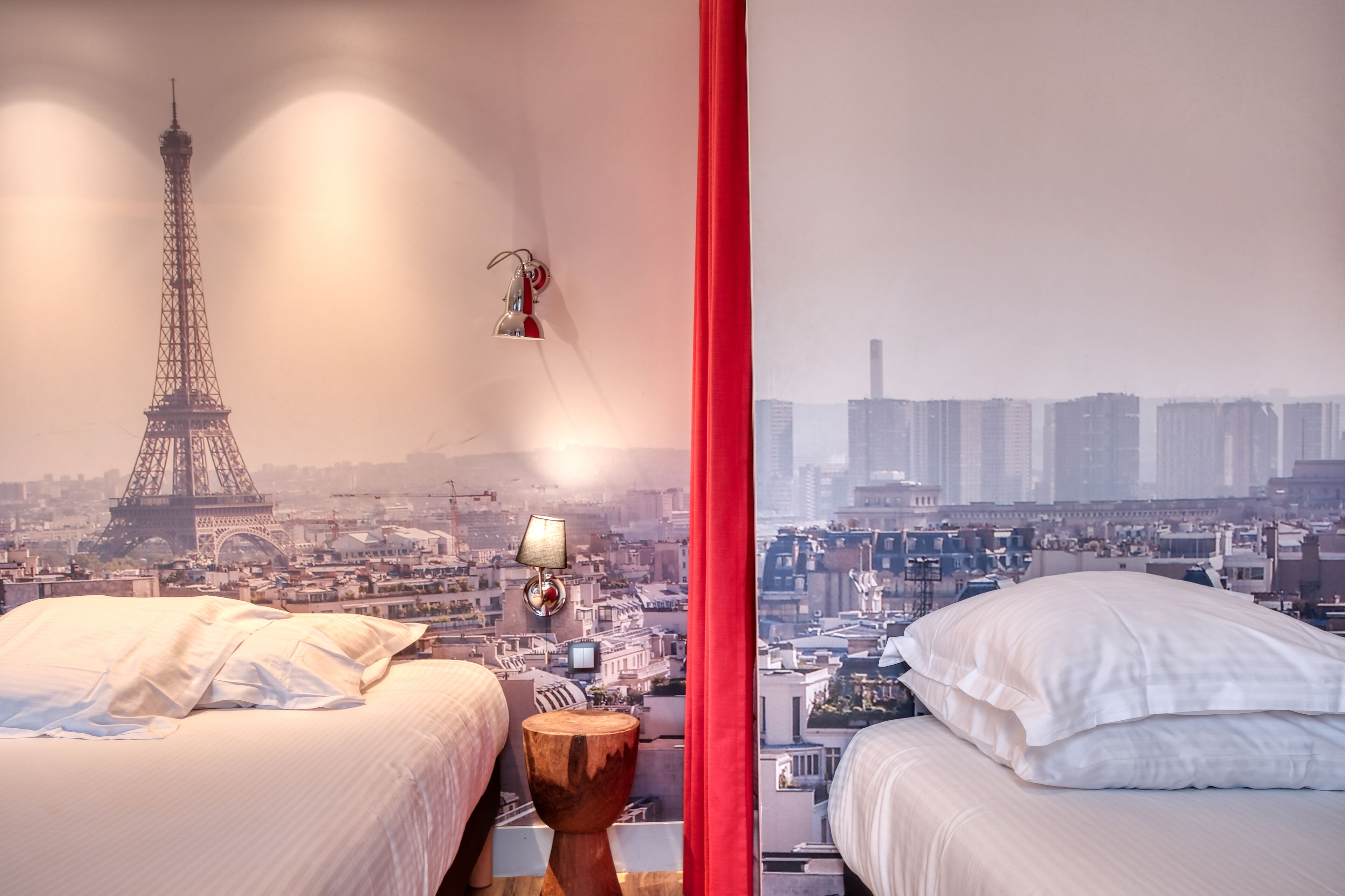 A hotel room with two beds, a wooden nightstand, a lamp, and a view of the Eiffel Tower and cityscape.