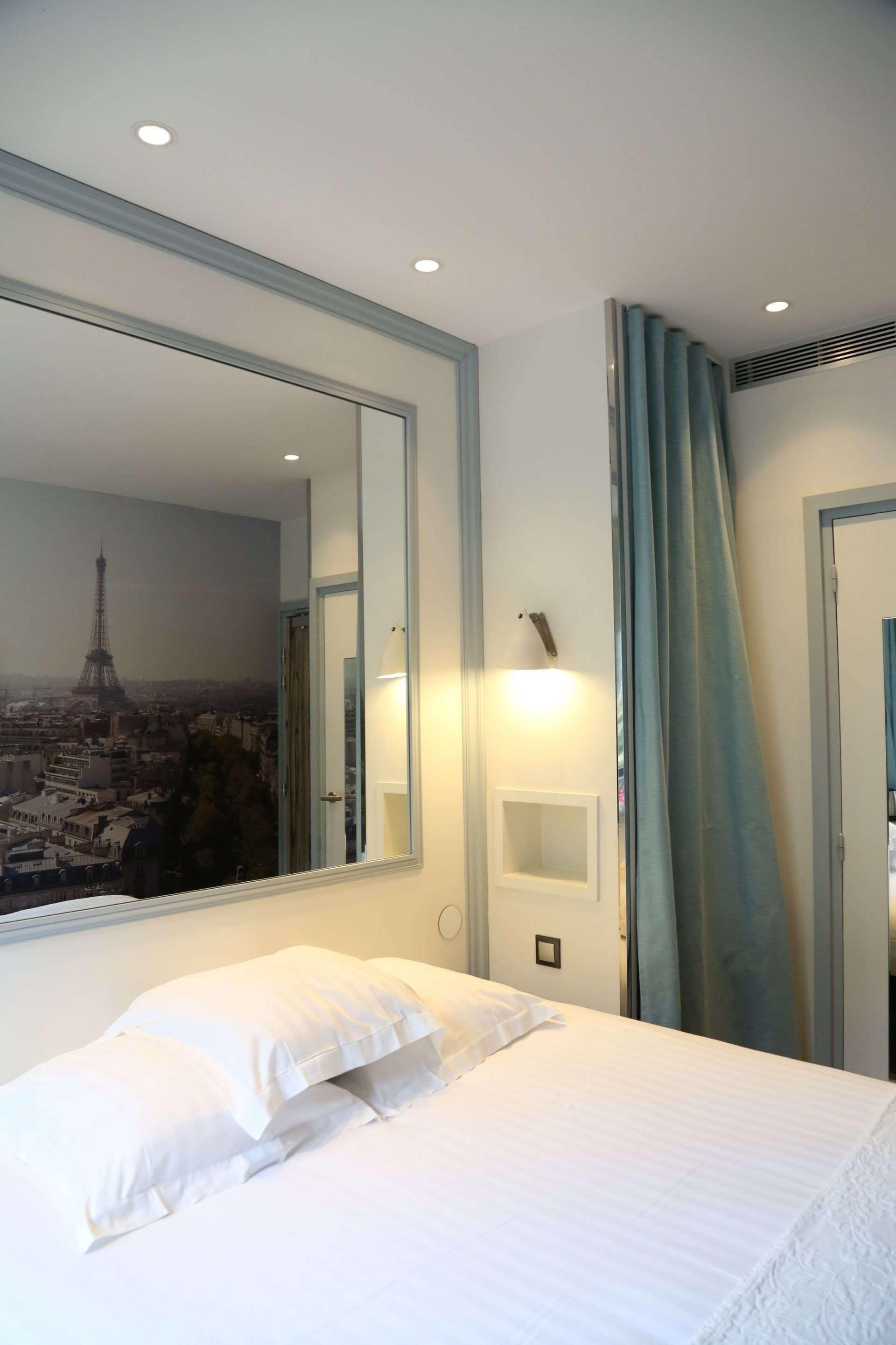A hotel room with a bed, a large mirror, a wall-mounted light, and a view of the Eiffel Tower through the window.