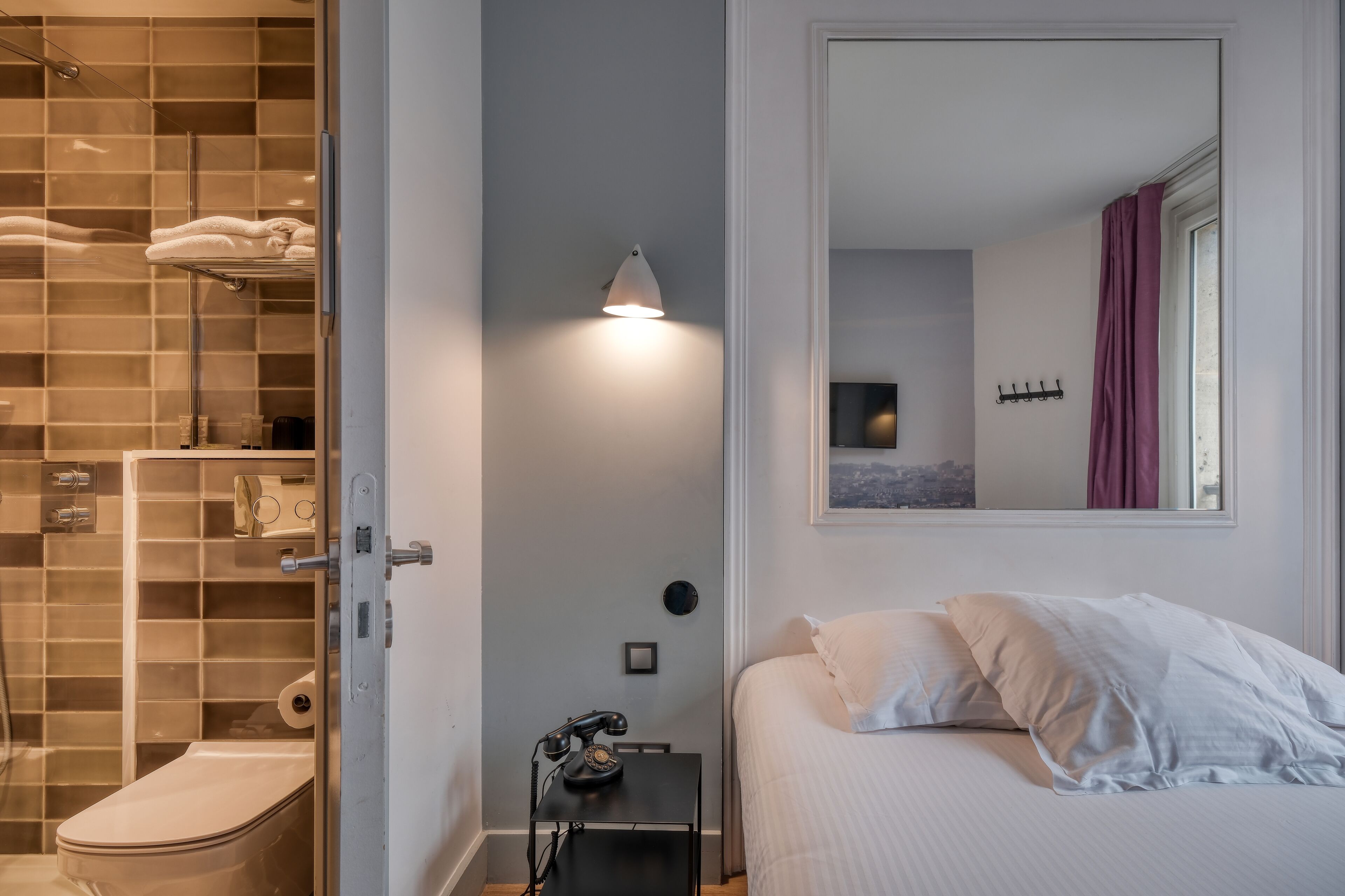 Et badeværelse med toilet, bruser og et håndklædestativ. Der er en seng med hvide sengetøj og et natbord med en lampe.