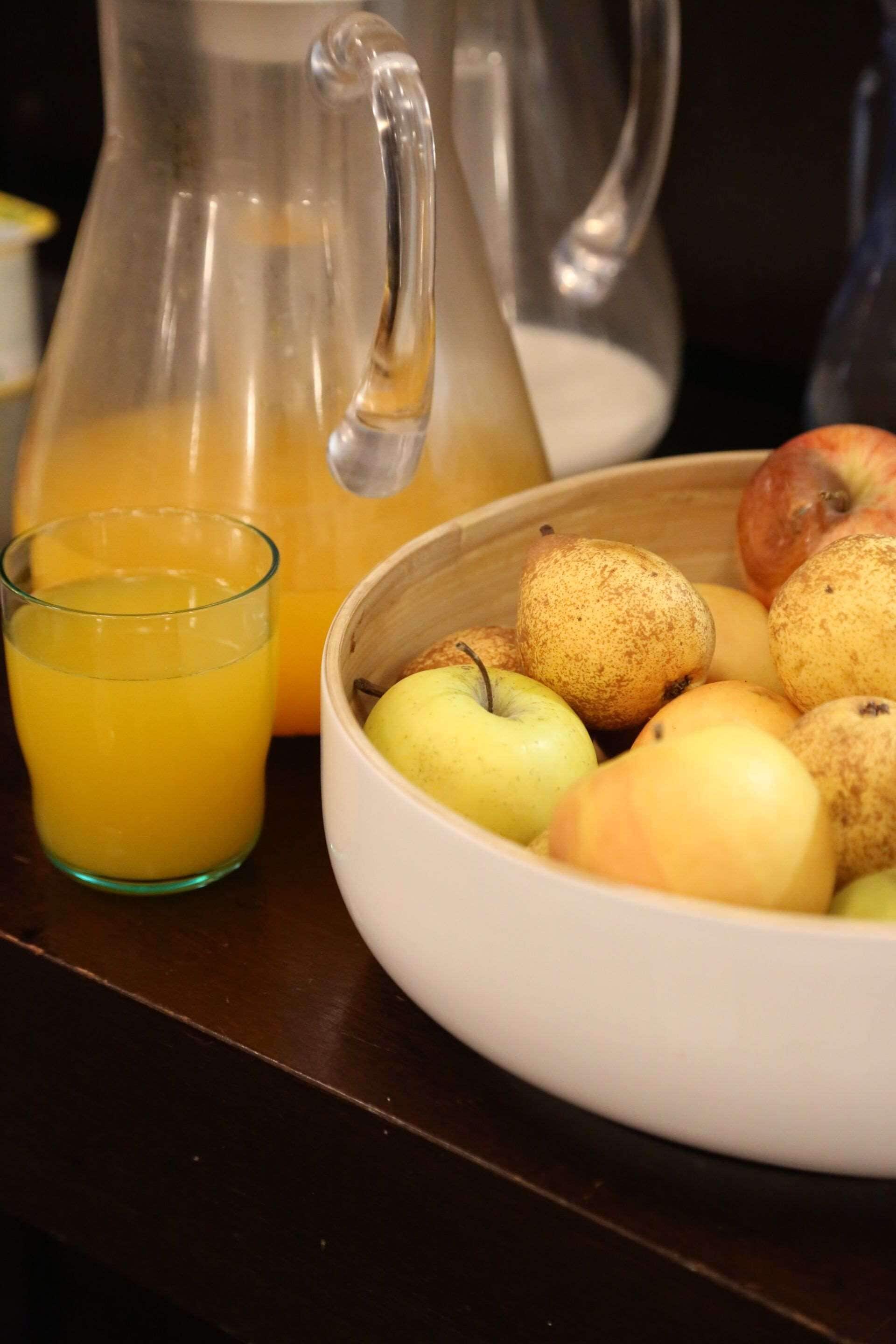 A pitcher of orange juice, a glass of orange juice, and a bowl of apples on a table.