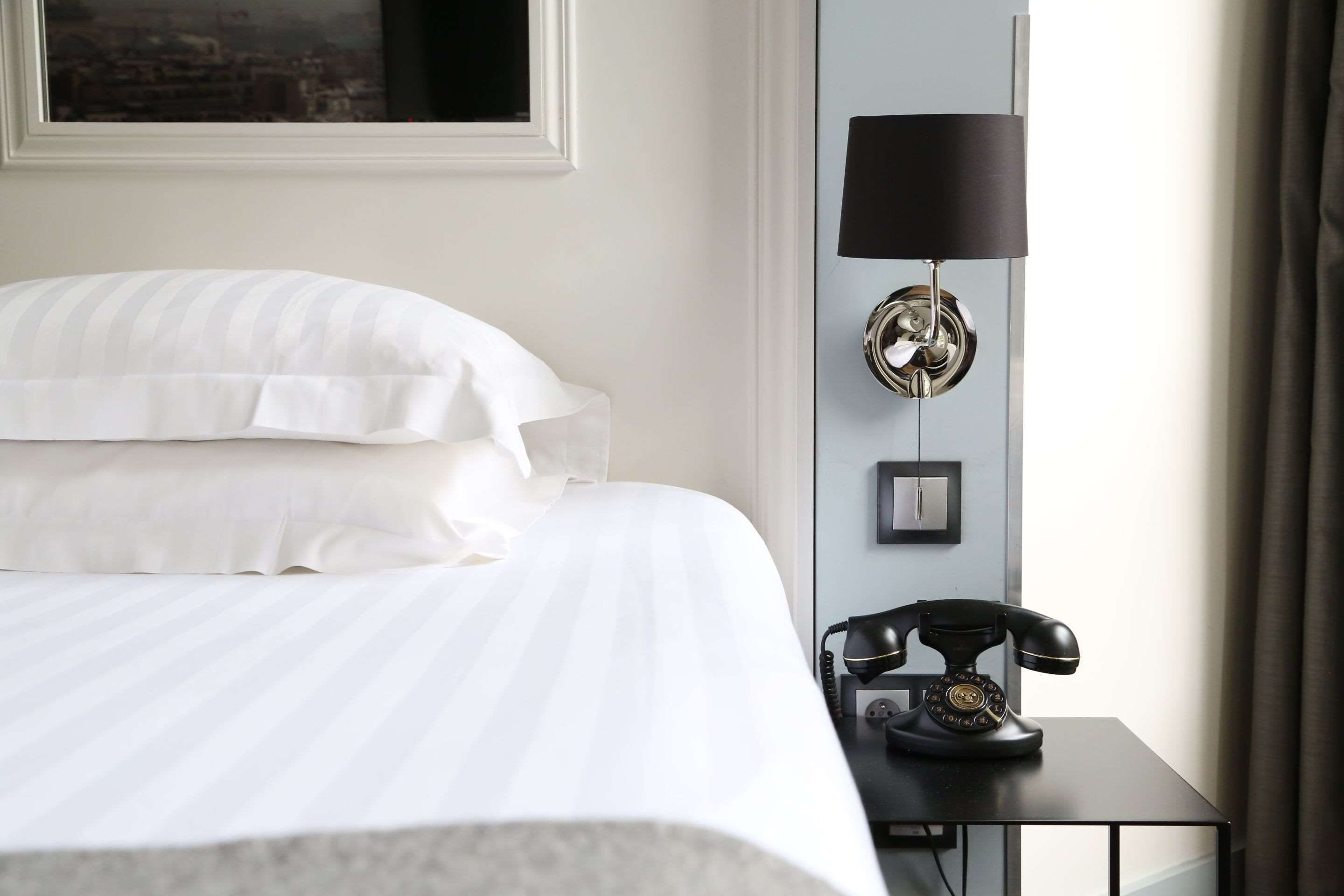 A neatly made bed with white linens, a black telephone on a small table beside it, and a wall-mounted lamp.