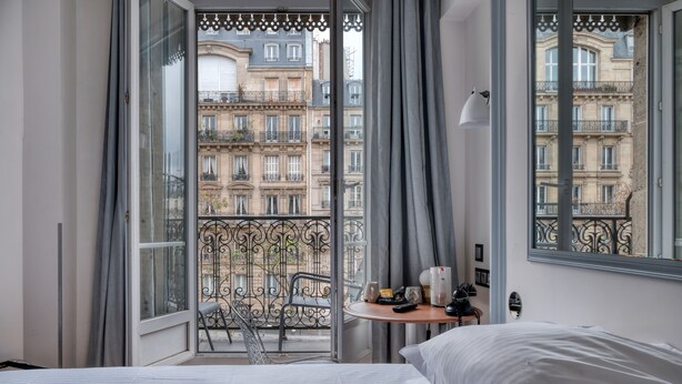 Et hotelværelse med en seng, et lille bord med en lampe, en stol og en balkon med udsigt til bygninger.