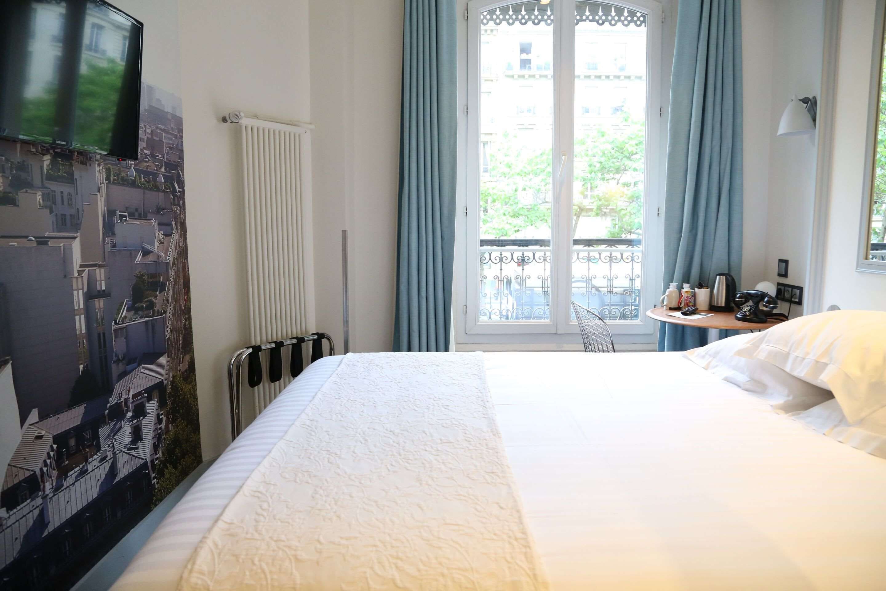 A hotel room with a large bed, a radiator, a window with curtains, a view of buildings, and a small table with items.