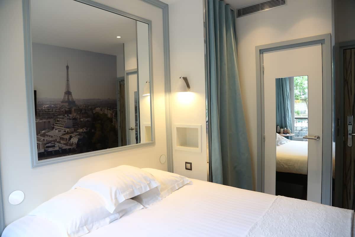 A hotel room with a bed, a large mirror, a wall-mounted light, and a view of the Eiffel Tower through the mirror.