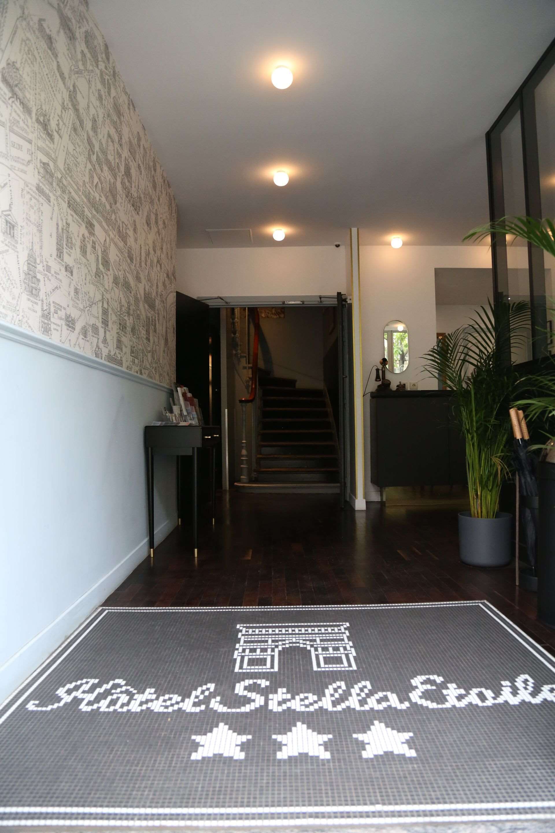 A hotel entrance with a dark wooden floor, a reception desk, a staircase, and a wall with a patterned design.