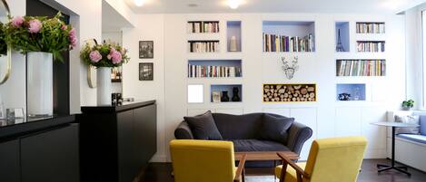Una sala moderna con un sofá oscuro, sillones amarillos, una mesa de centro y una estantería llena de libros y adornos.