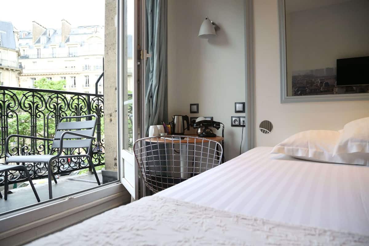 A hotel room with a bed, a chair, a desk with a lamp, and a view of the city through a window.