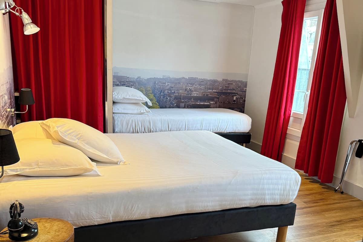 A hotel room with a large bed, a wooden side table, a lamp, and a city view through the window.