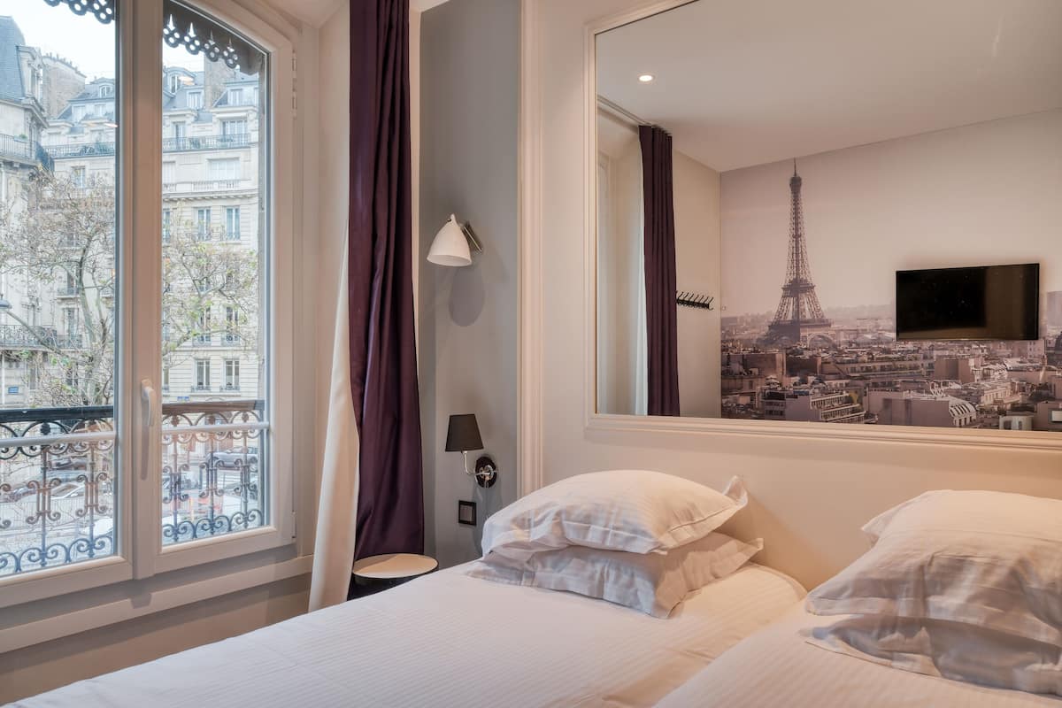 A hotel room with a bed, a window with curtains, a wall-mounted TV, and a cityscape mural.