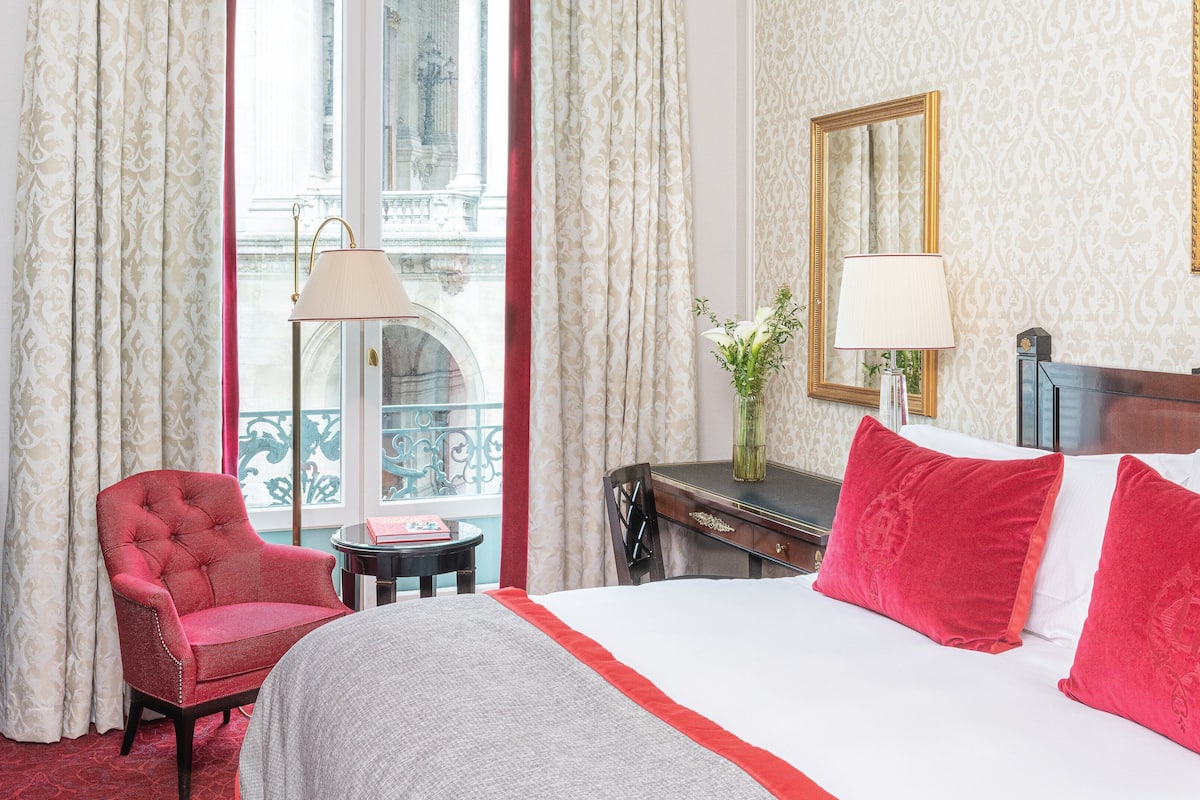 A hotel room with a bed, a red armchair, a small table with a lamp, and a view of a balcony with curtains.