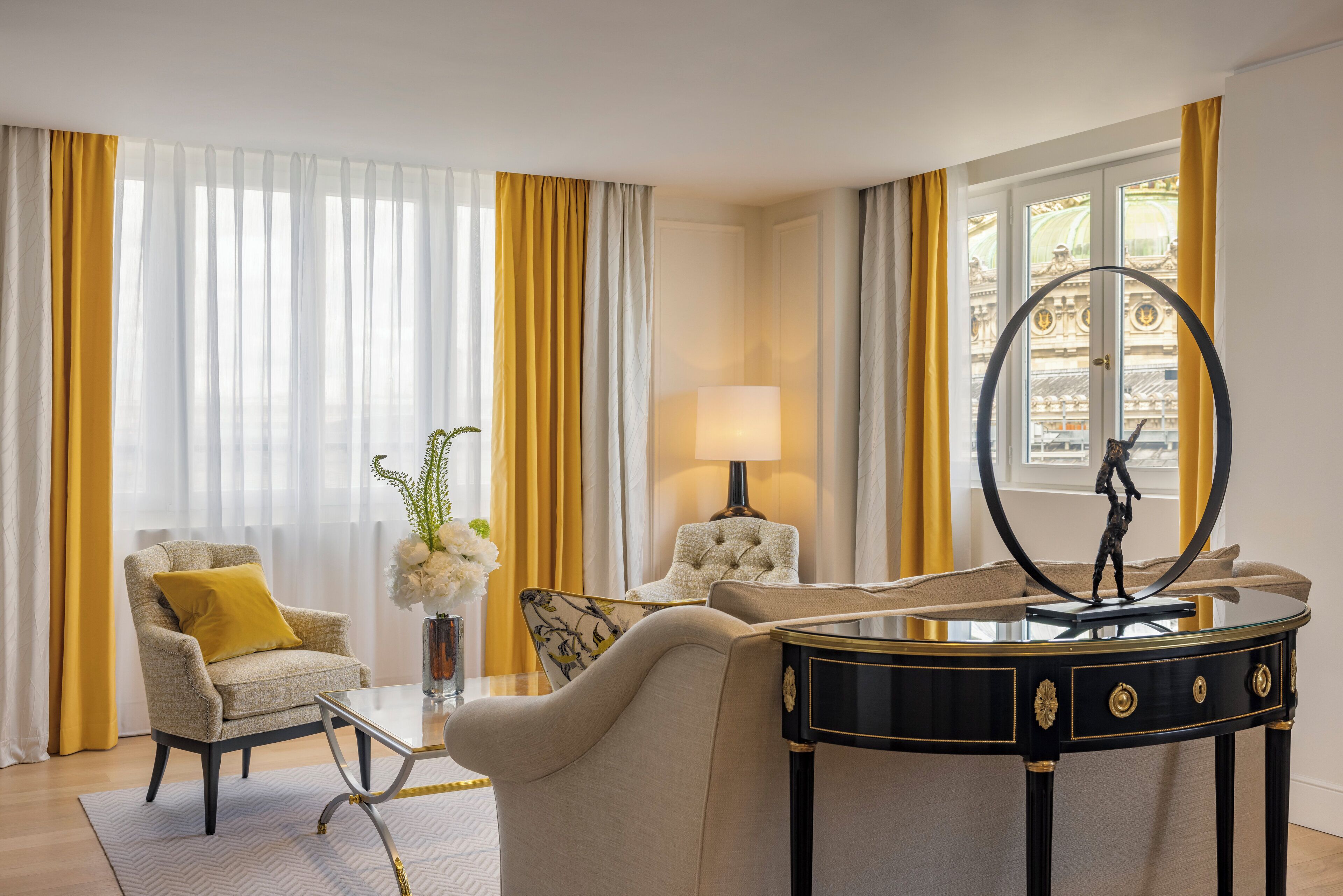 Un salón bien iluminado con un sofá, un sillón y una mesa de centro. Hay una lámpara sobre una mesita auxiliar y un espejo decorativo sobre un aparador.