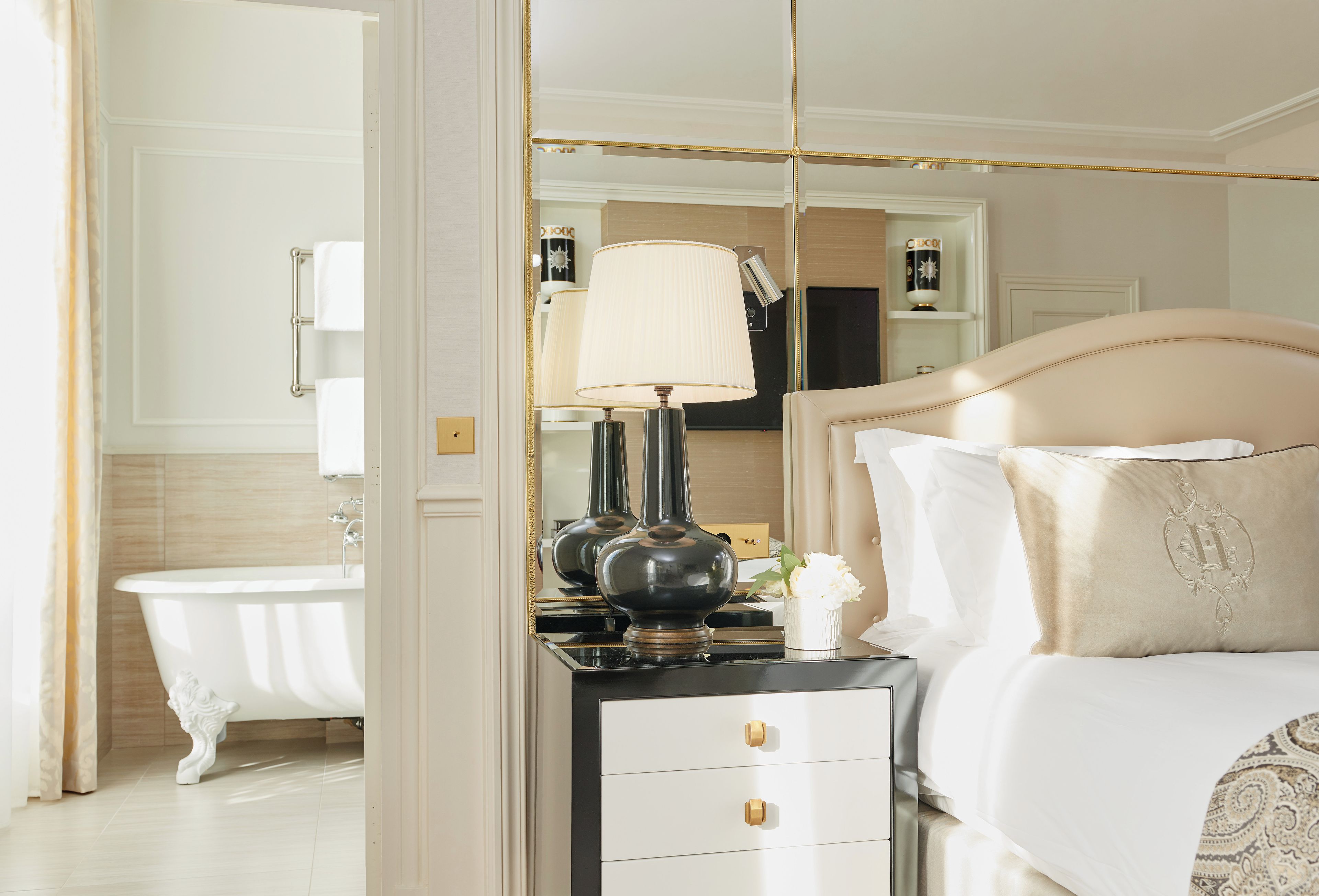 Habitación de hotel con cama, mesita de noche con lámpara, espejo y baño con bañera.