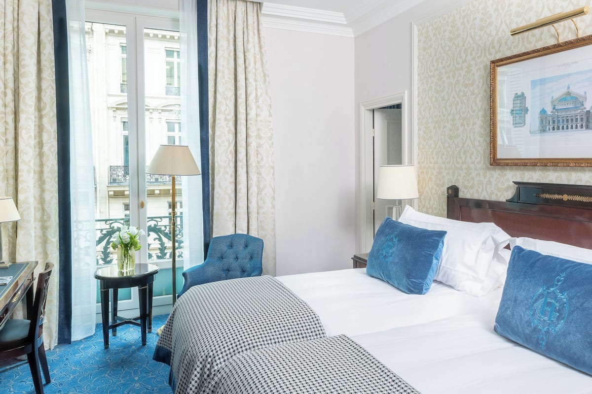 A hotel room with a large bed, a chair, a small table with a lamp, and a view of a building through the window.