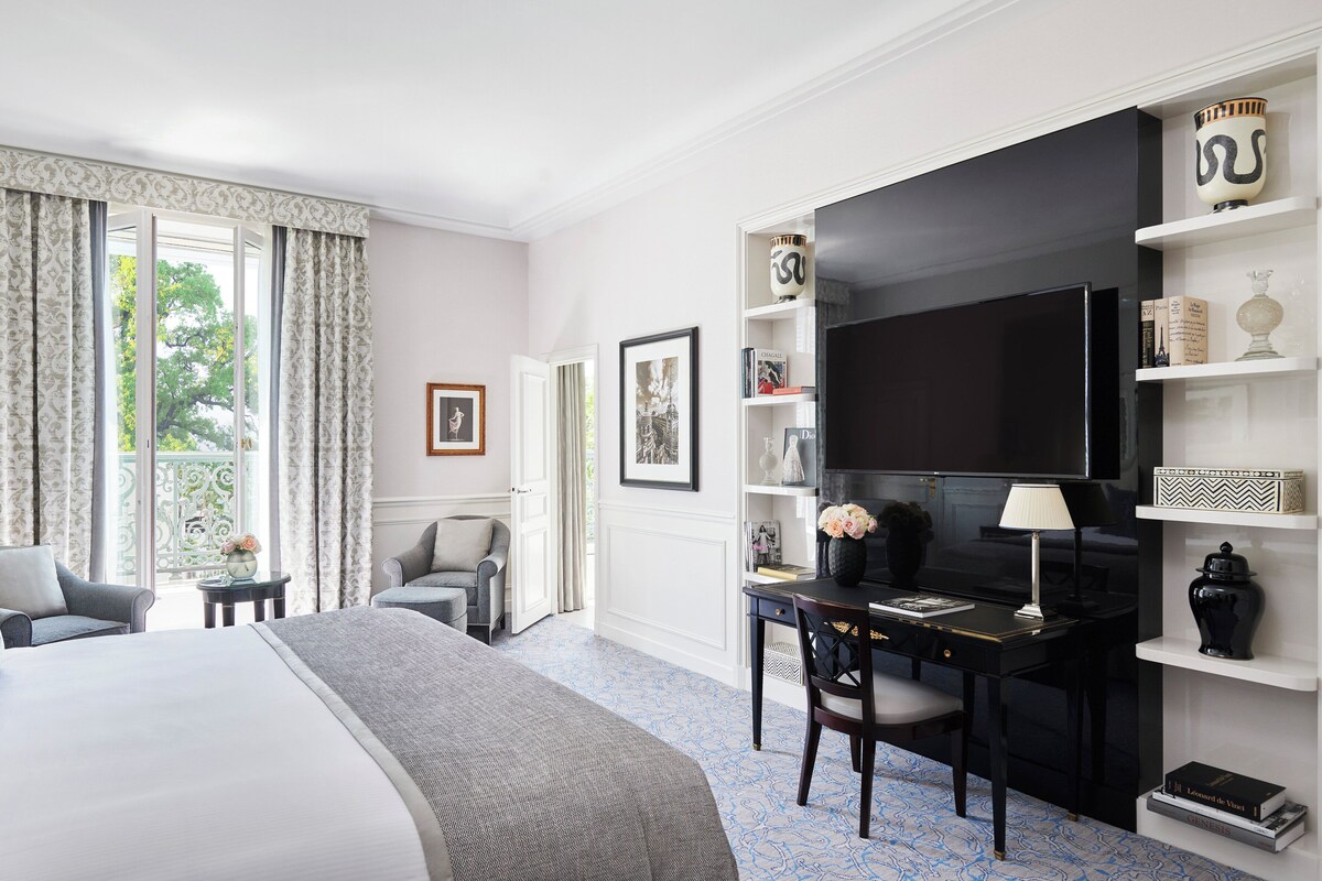 A hotel room with a bed, a desk, a chair, a TV, and a bookshelf.