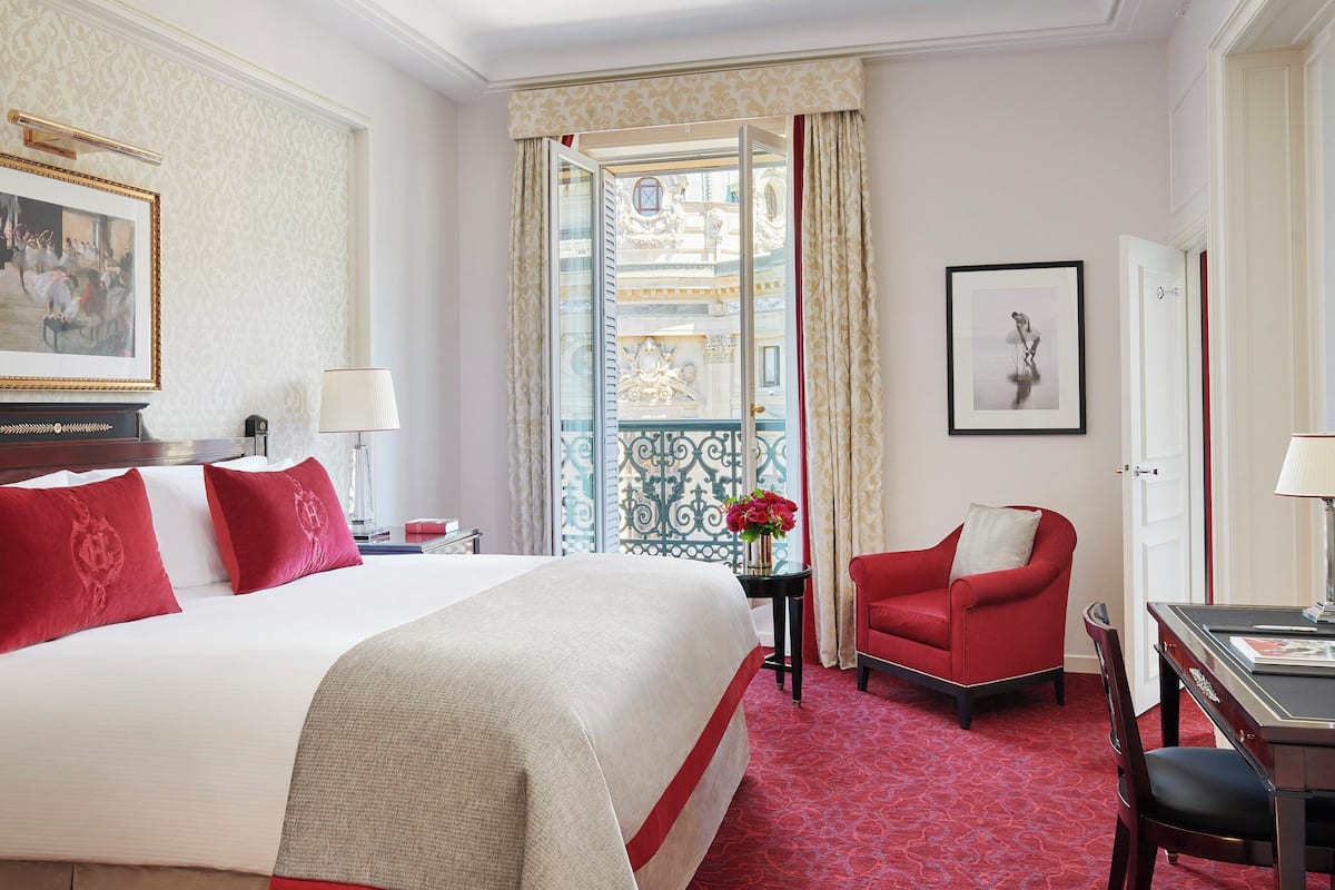 A hotel room with a large bed, a desk, a chair, a nightstand, a lamp, a framed picture, and a view of a balcony.