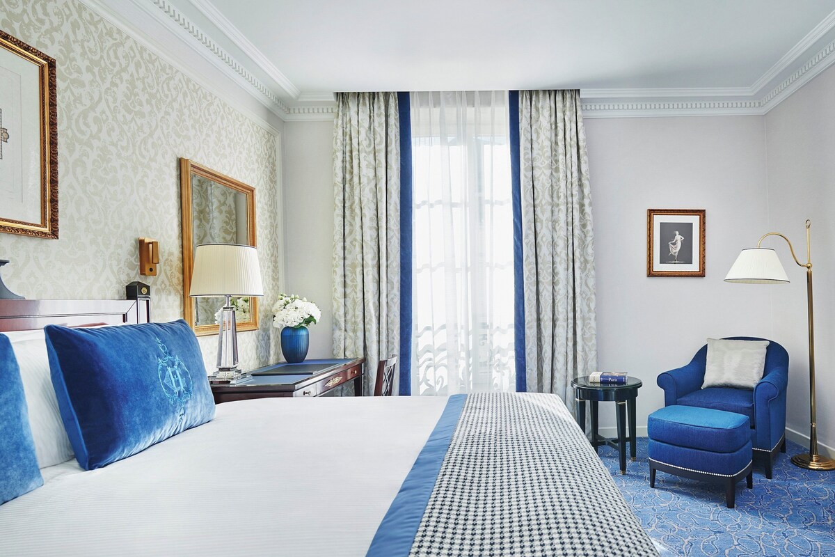 A hotel room with a large bed, a desk with a lamp, a chair, a television, and a window with curtains.