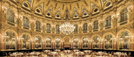 A grand ballroom with ornate chandeliers, multiple round tables, and a central fireplace.