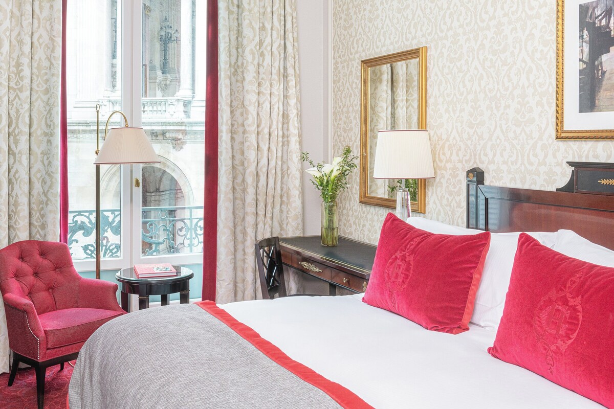 A hotel room with a bed, a red armchair, a small table with a lamp, and a view of a balcony with curtains.