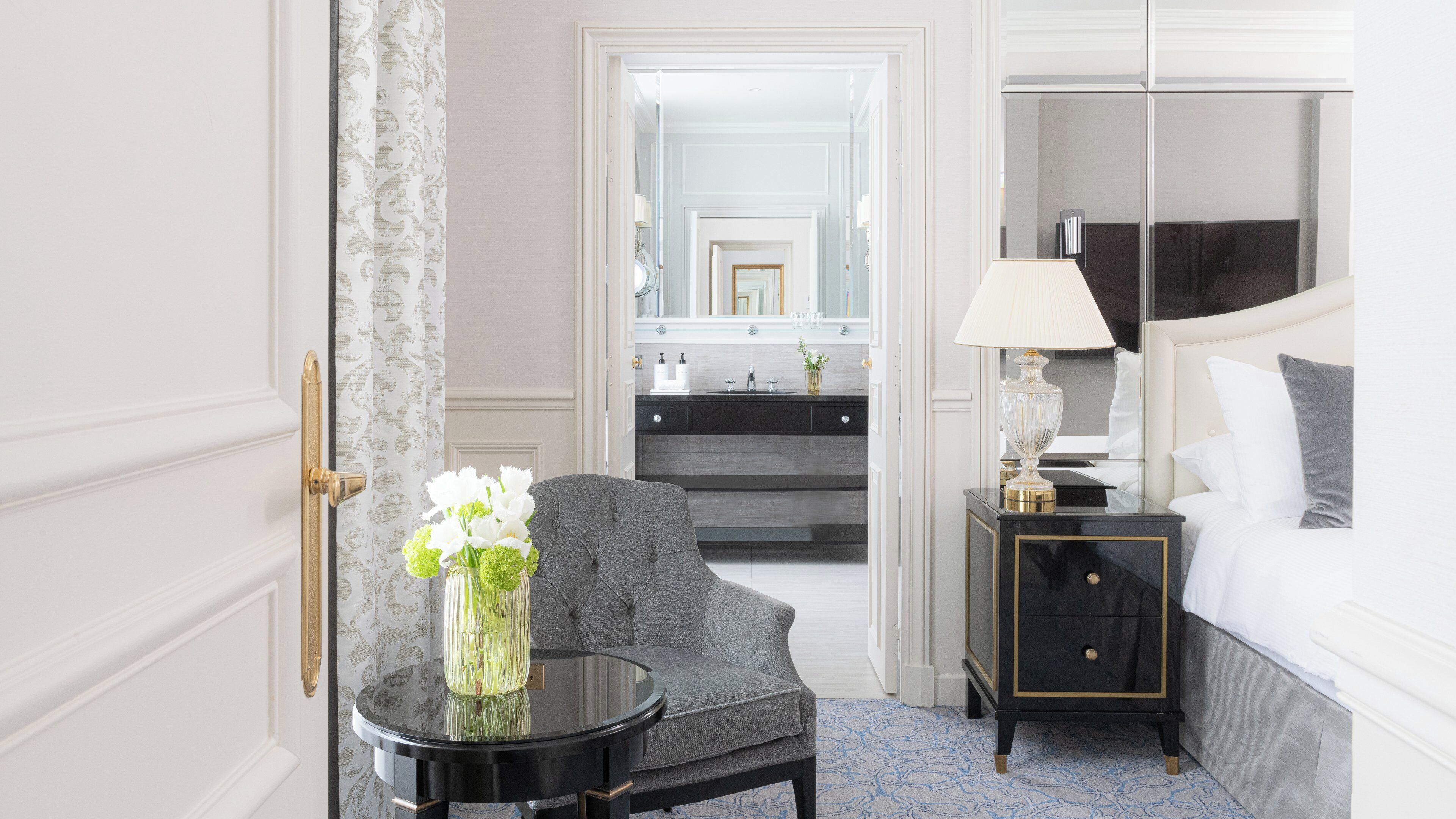 A hotel room with a bed, a nightstand, a chair, a round table with a vase of flowers, a mirror, and a bathroom visible through an open door.