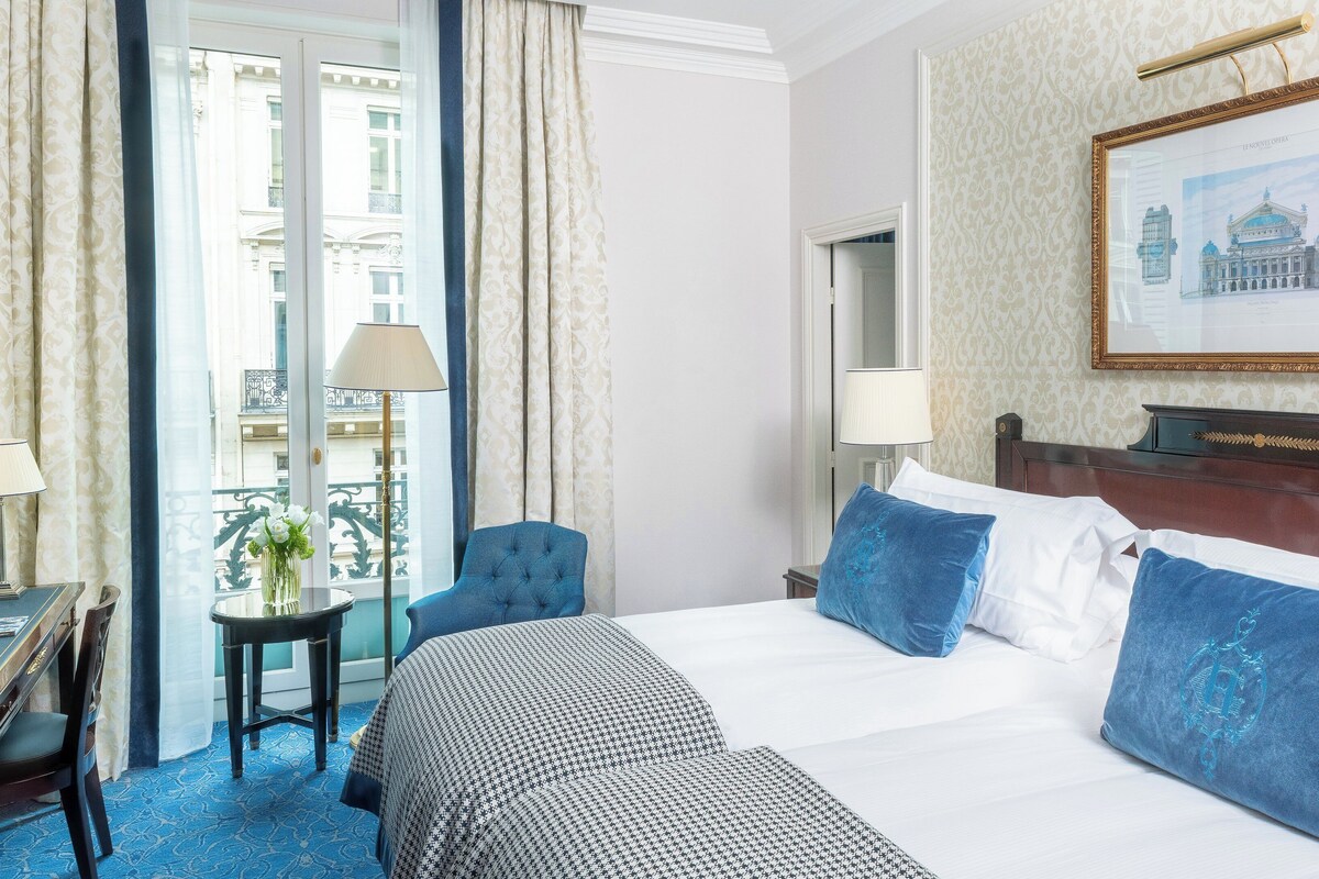 A hotel room with a large bed, a chair, a small table with a lamp, and a view of a building through the window.